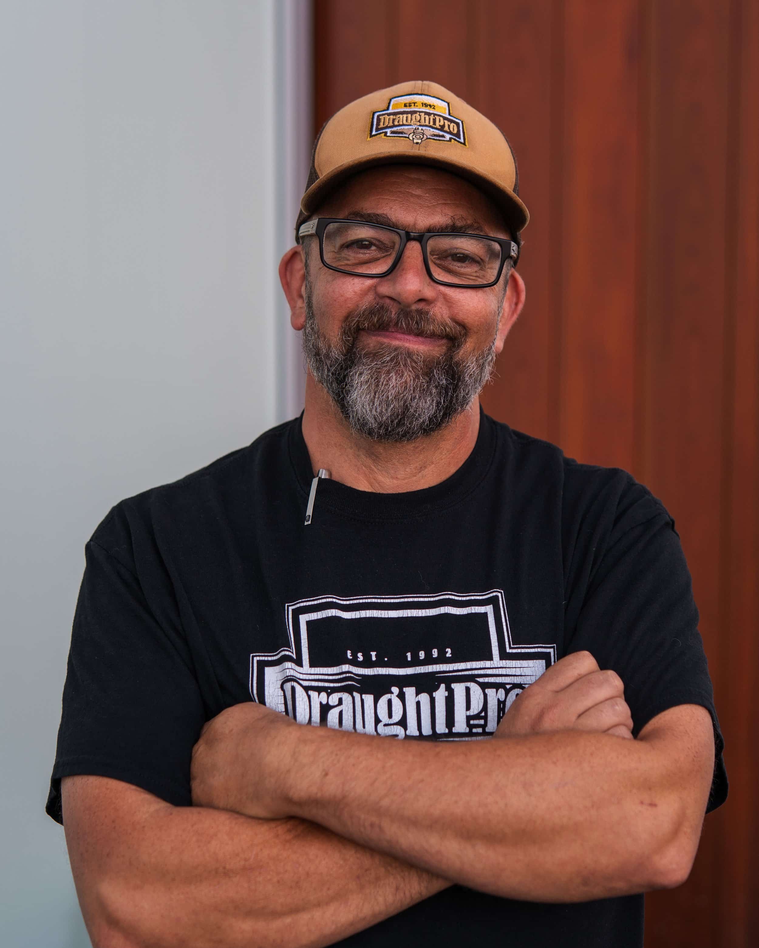A  man with glasses, a beard, and a mustache, wearing a tan baseball cap that says 'DraughtPro' and a black T-shirt with the same logo, standing with arms crossed in front of a background with wood paneling.