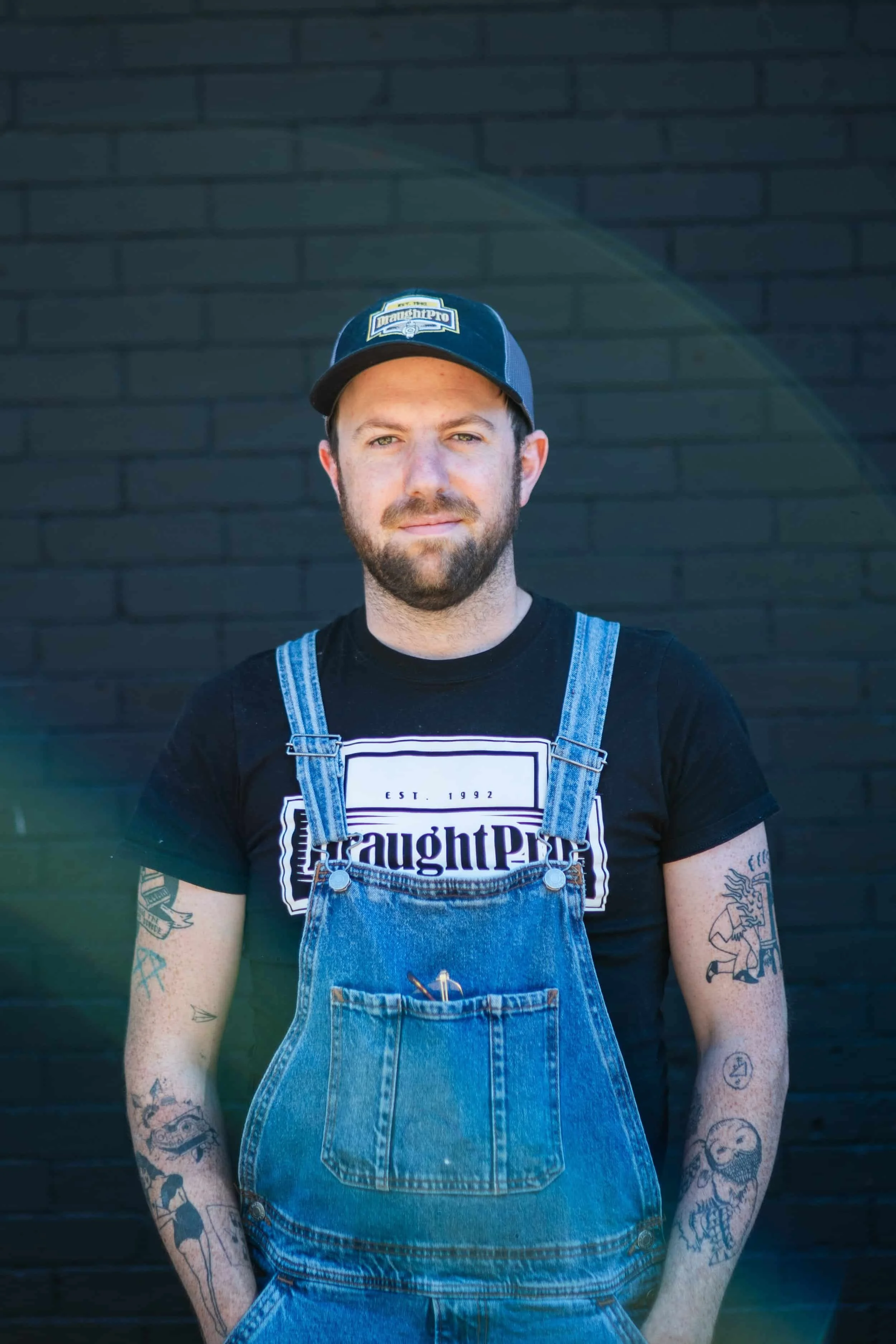 A man with a beard and tattoos wearing a black 'ThoughtP' t-shirt, denim overalls, and a blue baseball cap standing in front of a black brick wall.