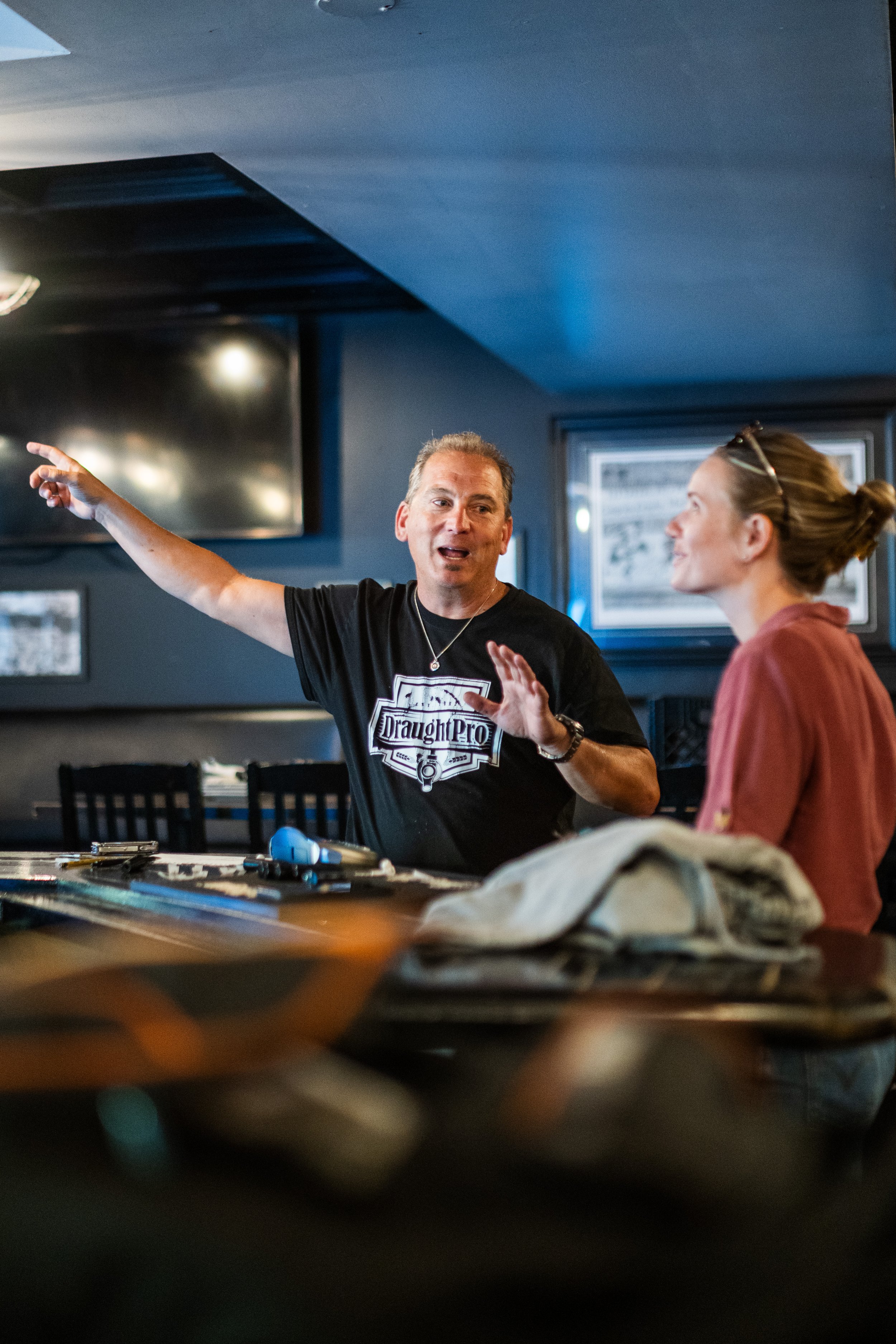 A man and woman are engaged in a conversation at a bar or restaurant, with the man animatedly gesturing with his arm extended and the woman listening attentively. The man is wearing a black T-shirt with a 'DraughtPro' logo, and the woman is wearing glasses and a rust-colored jacket.