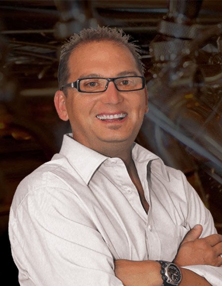 Smiling man with glasses wearing a white shirt, standing in an industrial or rustic setting.