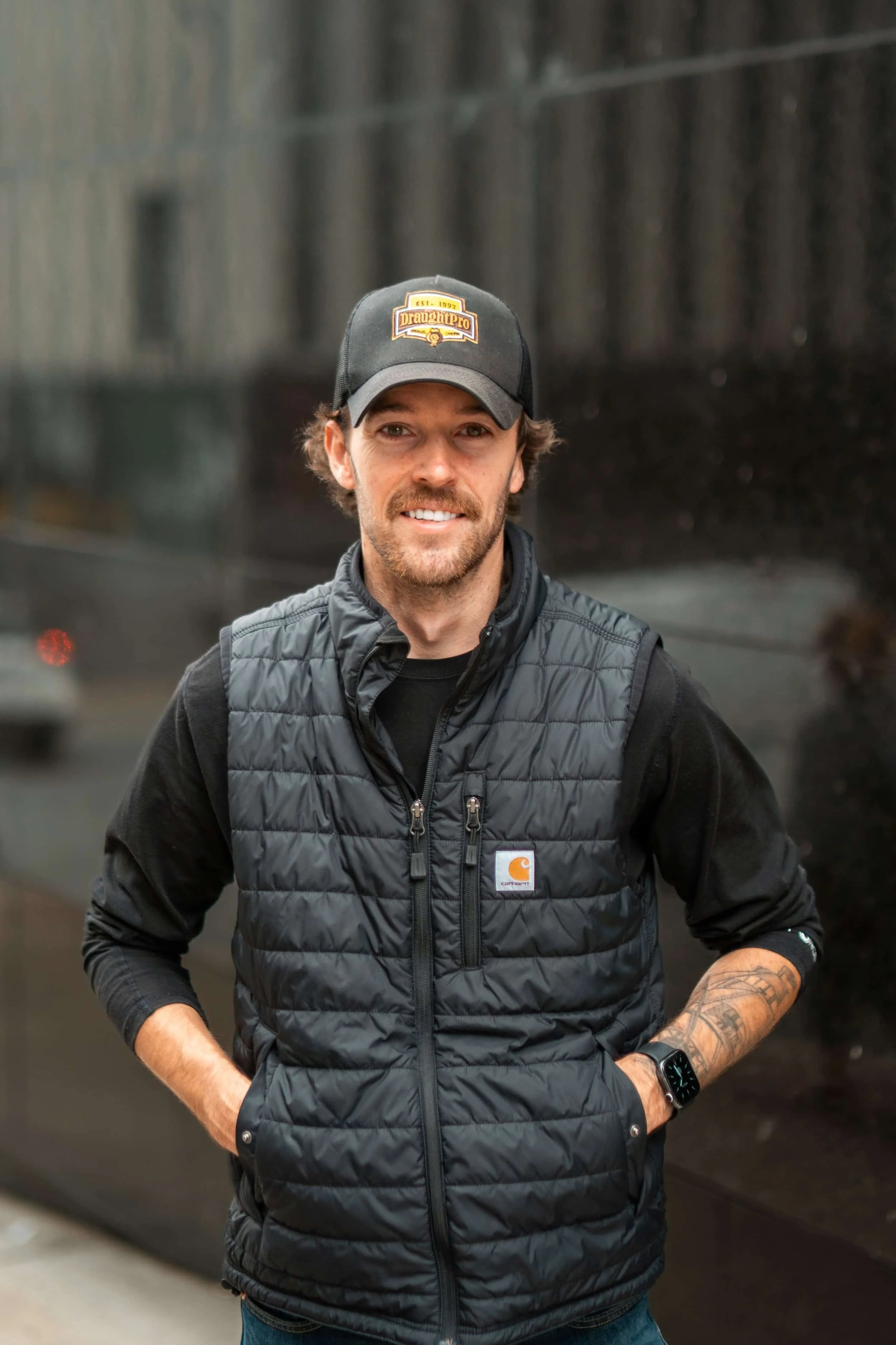 A man with brown hair, a beard, and tattoos on his left arm, wearing a black Carhartt vest, black shirt, and a black baseball cap with a yellow logo, stands outdoors with his hands in his pockets, smiling in front of a reflective black wall.