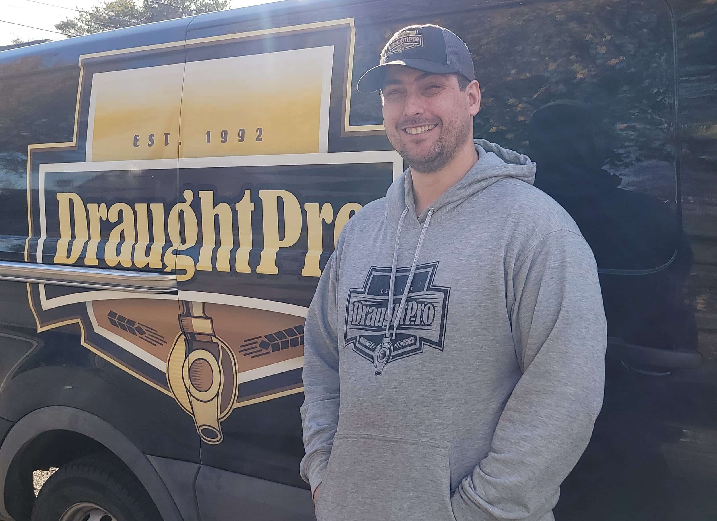 A smiling man wearing a gray hoodie and cap standing in front of a black vehicle with a logo and text that says 'Draught Pro' and 'Est. 1992'.