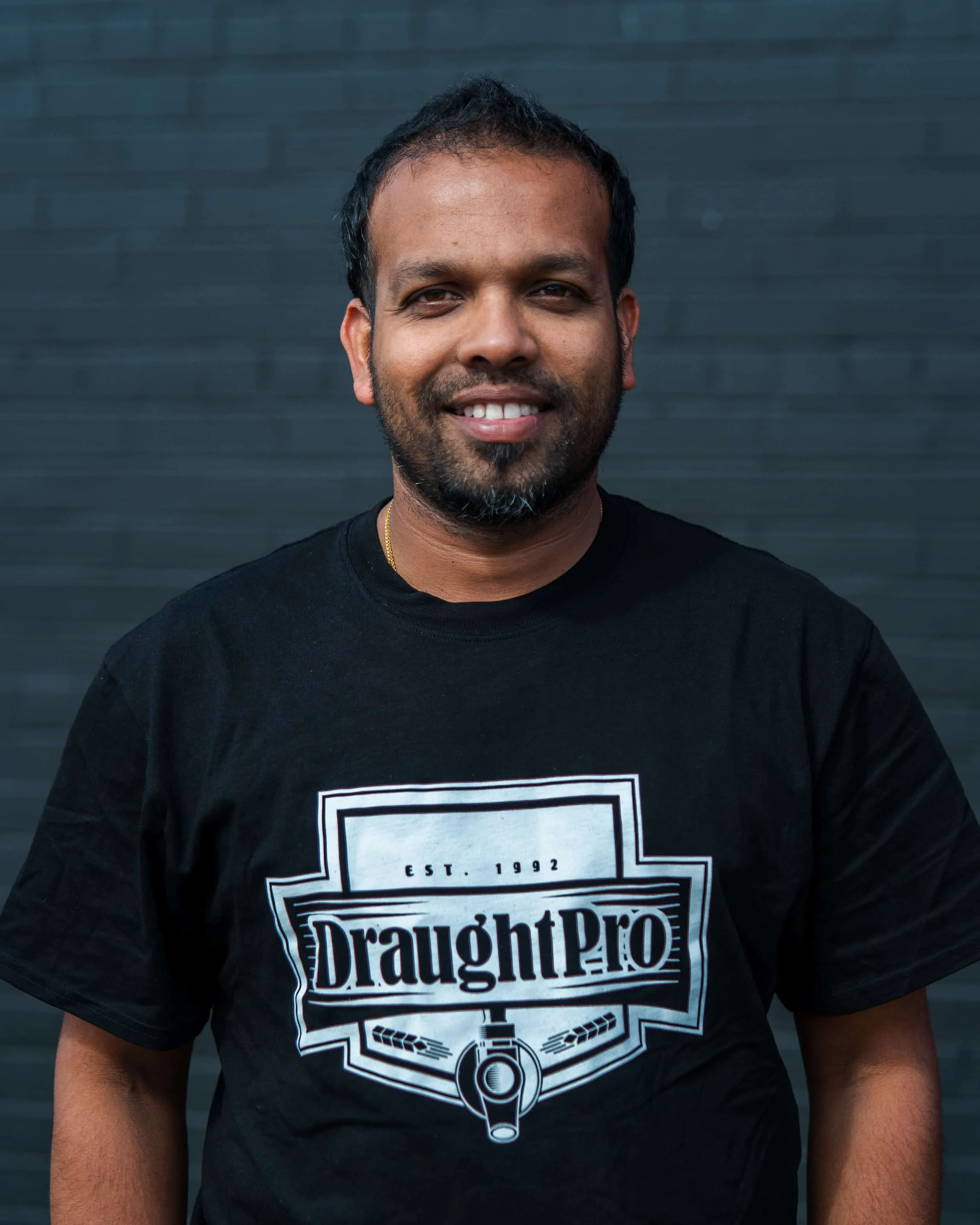 A man with short black hair and a beard, smiling, wearing a black t-shirt with a "DraughtPro" logo, standing in front of a dark gray wall.