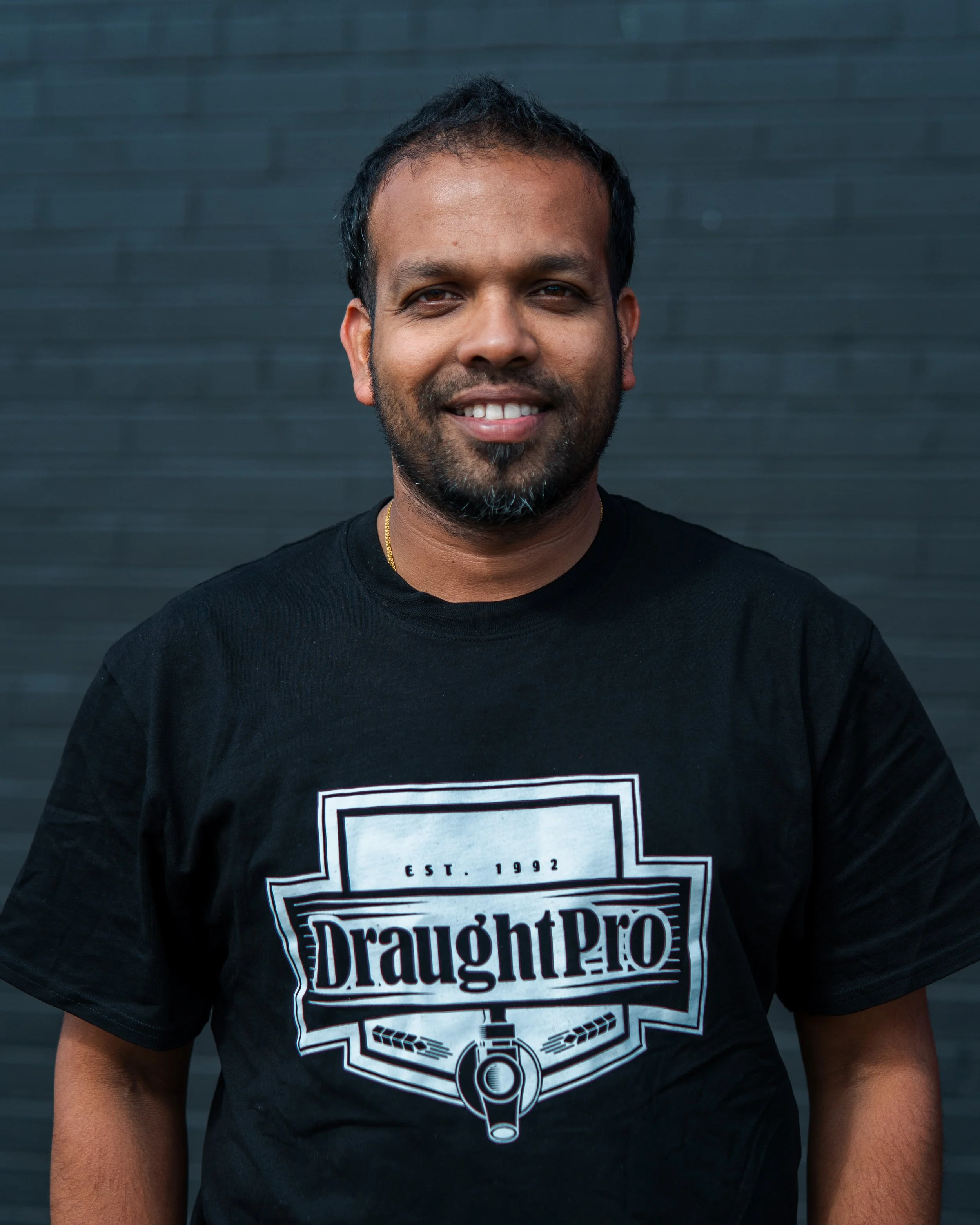 A man with short black hair and a beard, smiling, wearing a black t-shirt with a "DraughtPro" logo, standing in front of a dark gray wall.