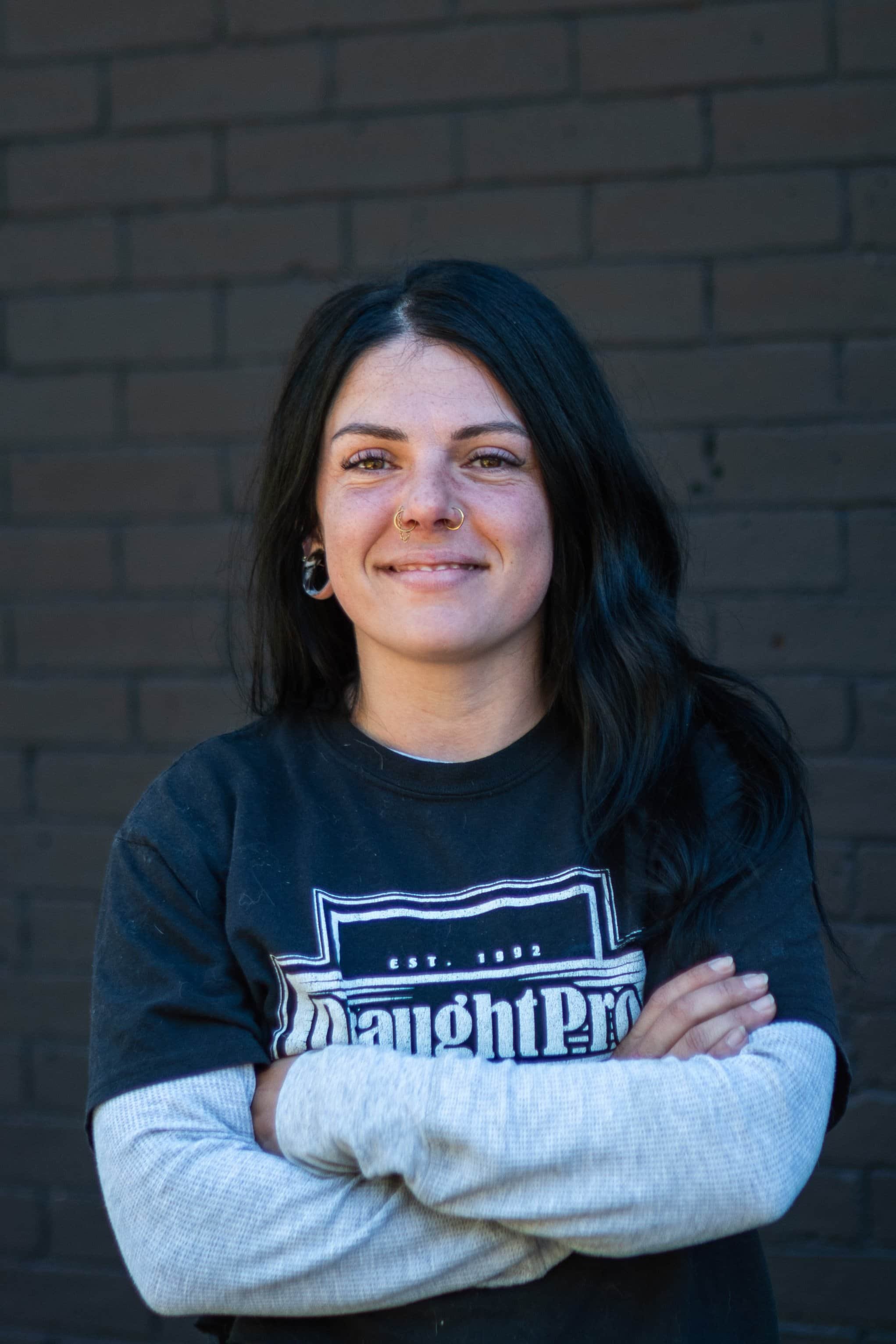 A woman with long dark hair, tattoos, and multiple piercings, smiling with crossed arms, standing against a black brick wall.