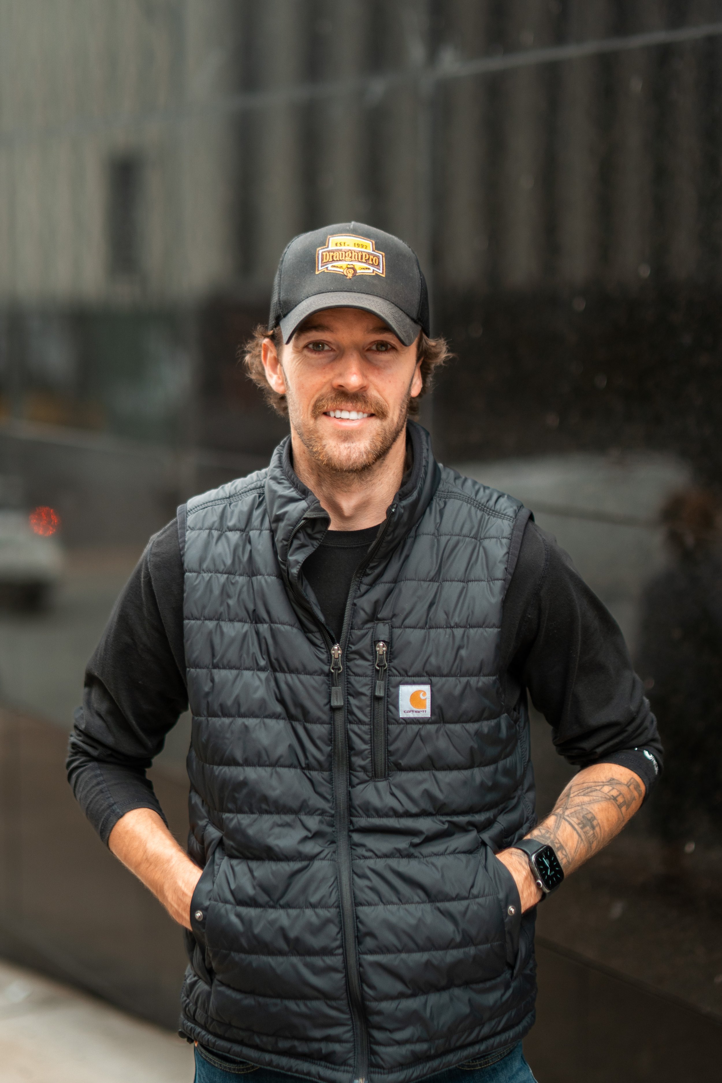 A man with a beard and tattoos wearing a black Carhartt vest, black long-sleeve shirt, black cap, and a watch, standing outdoors in front of a dark reflective surface.