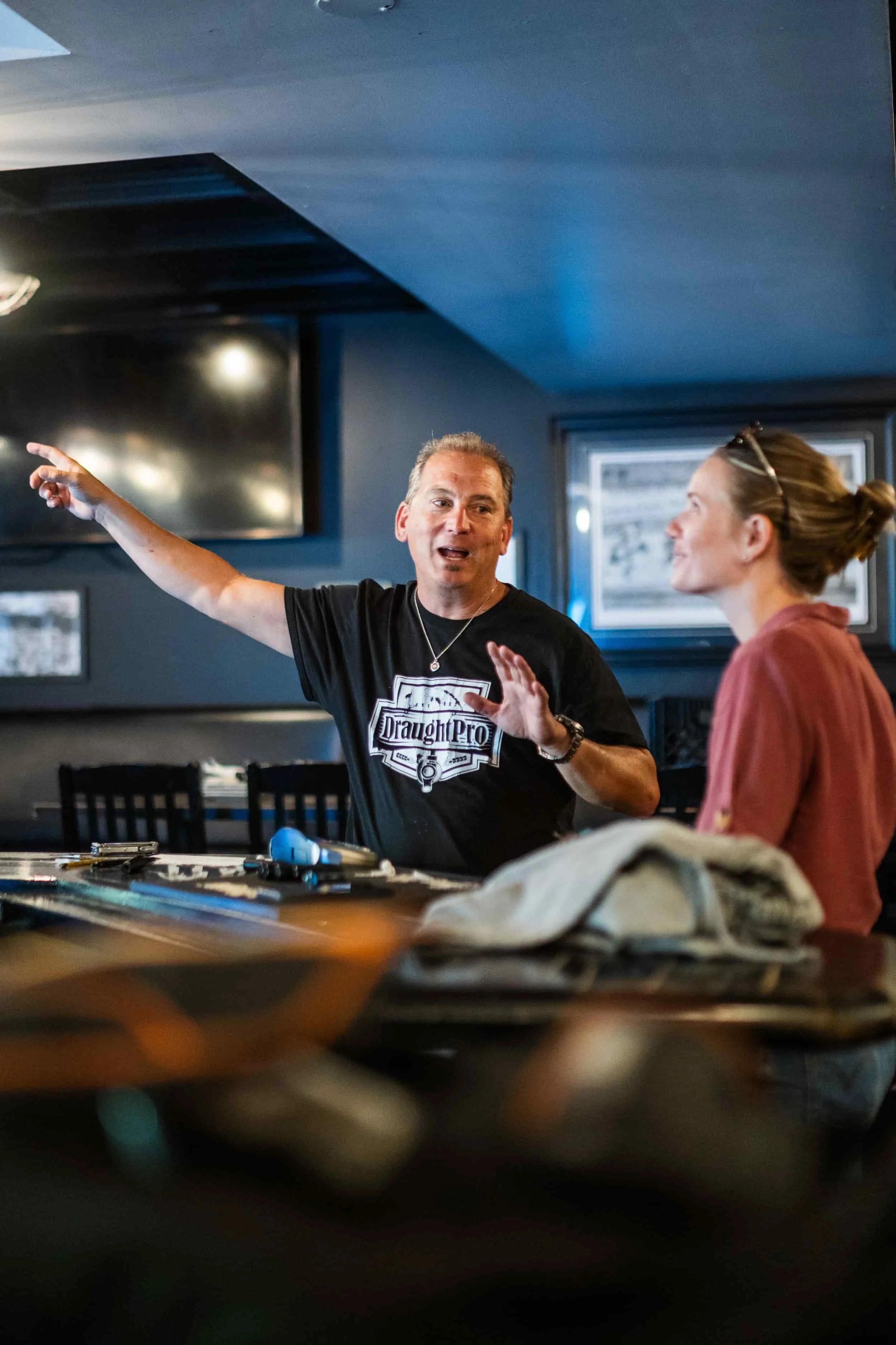 Man in black t-shirt gesturing while talking to woman in glasses and red shirt inside a dimly lit room with framed pictures on the wall.