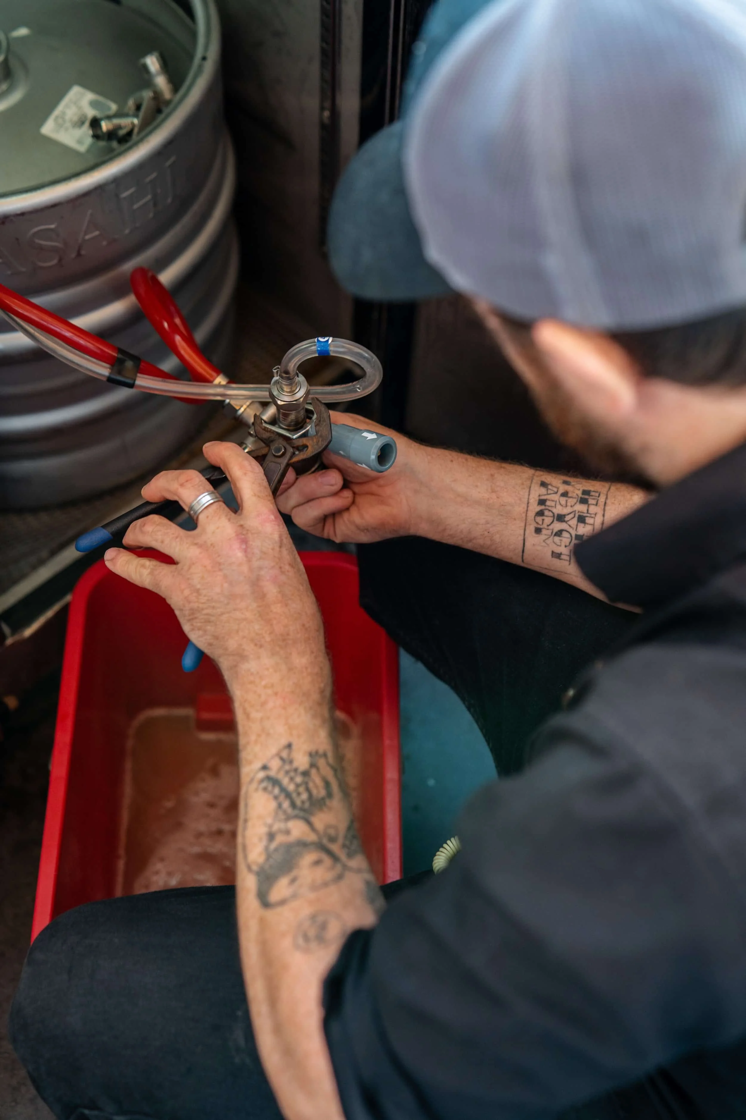 A person with tattoos on their arms and wearing a baseball cap is connecting beer gas to a keg in a bar setting.