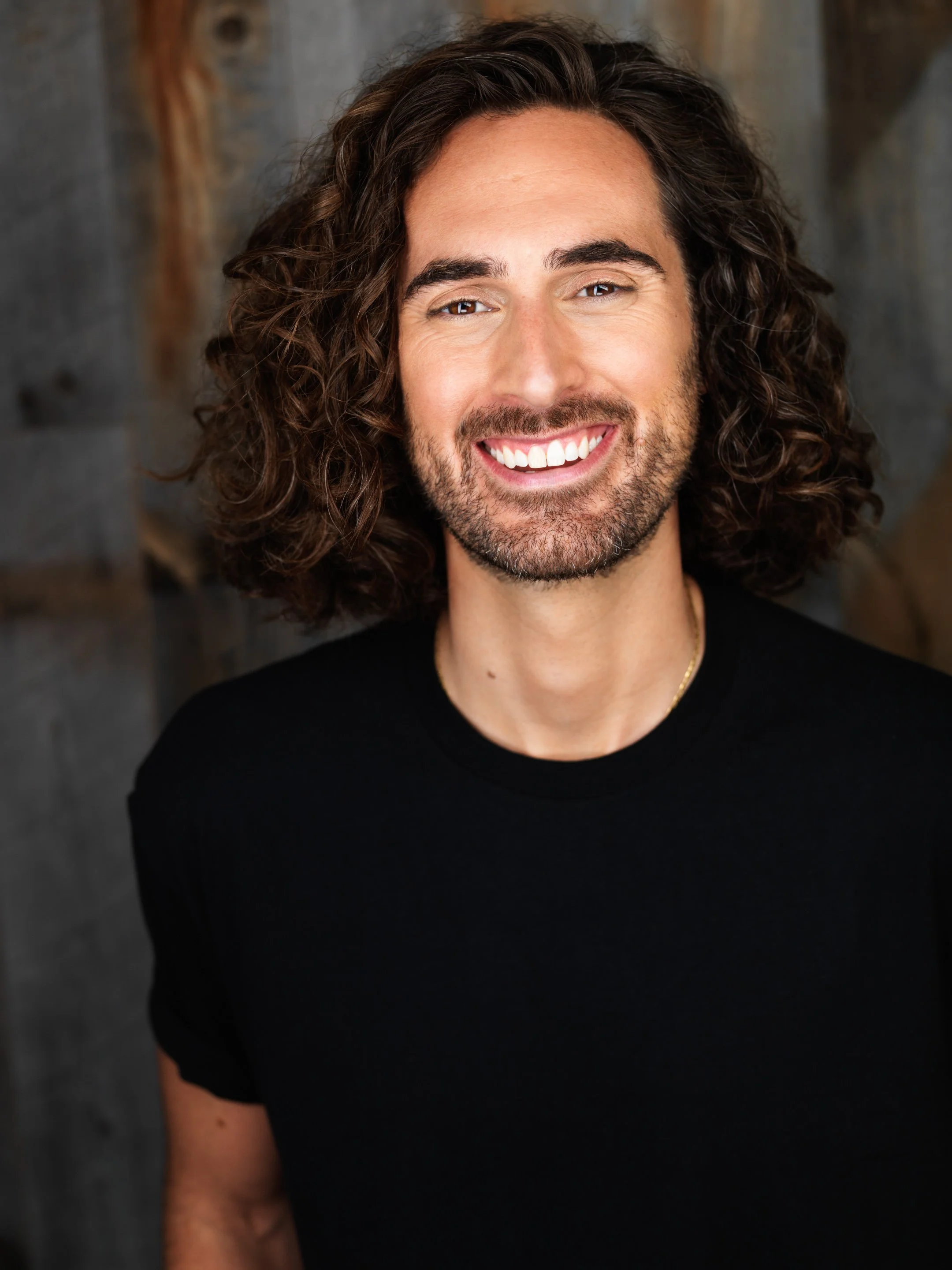 A smiling man with long curly hair and a beard wearing a black shirt, standing against a wooden background.