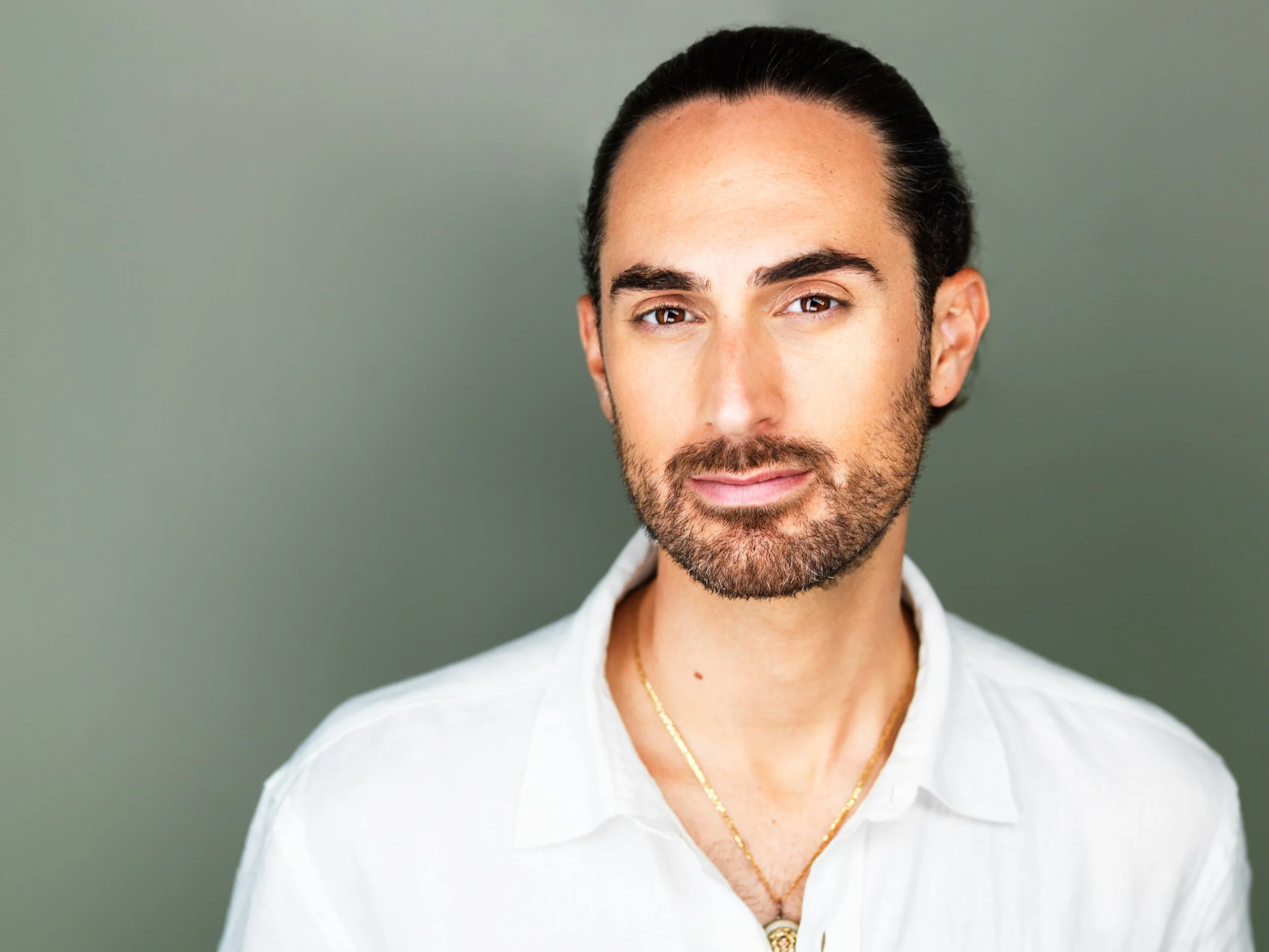 Close-up portrait of a man with dark hair and beard wearing a white shirt and gold chain, standing against a plain green background.