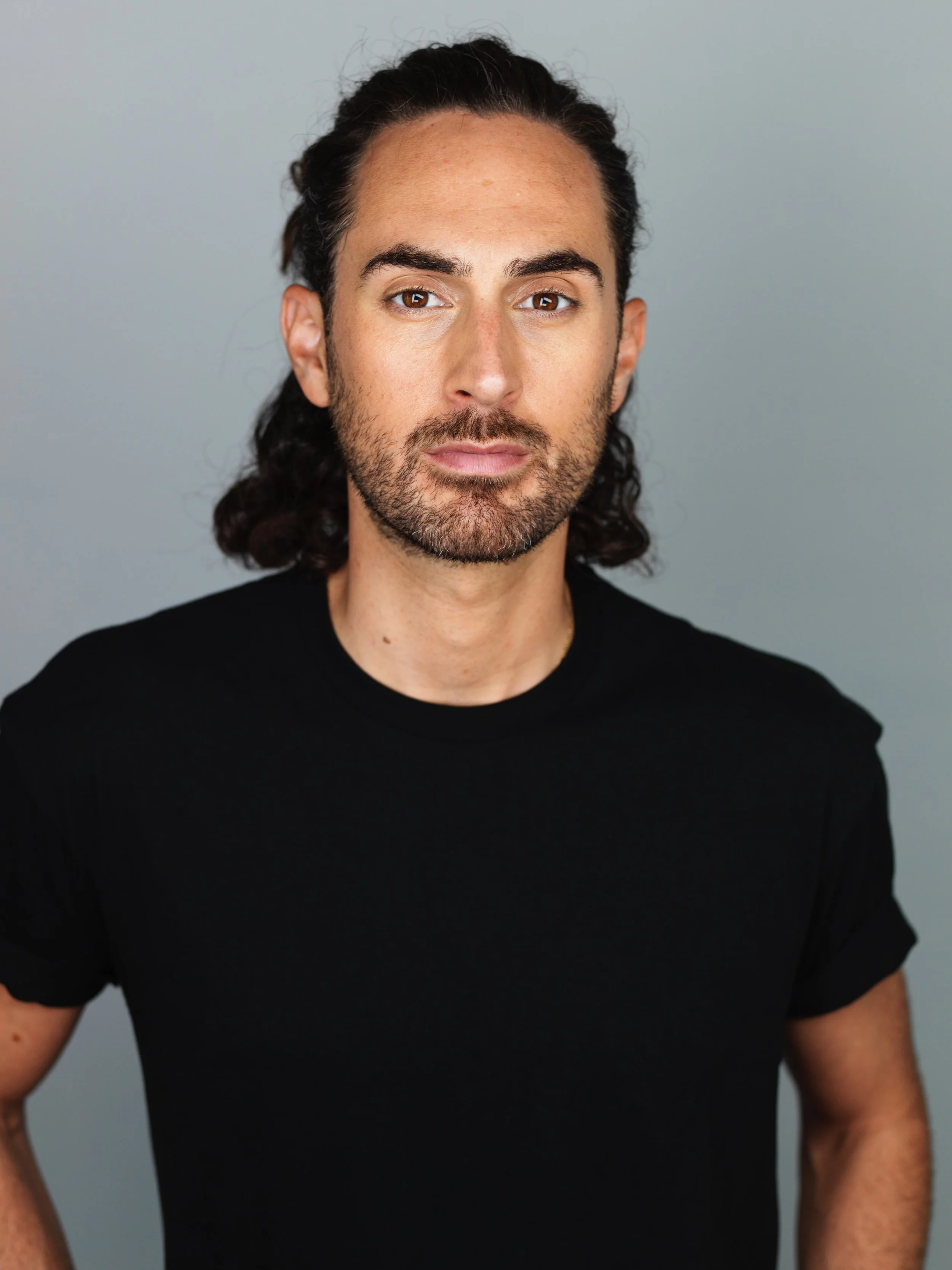 A man with long dark hair, a beard, and wearing a black t-shirt, standing against a neutral background.