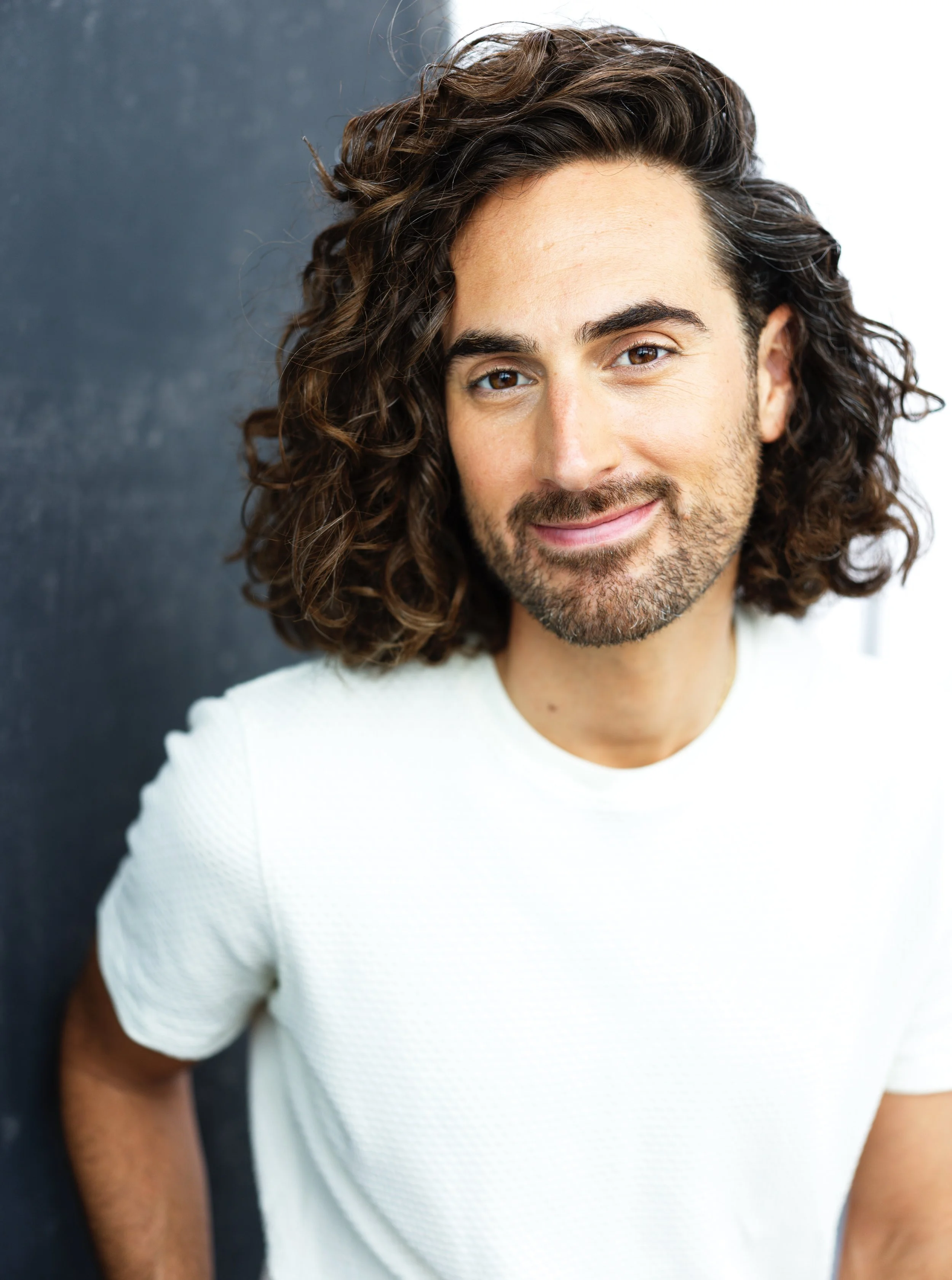 Close-up of a smiling man with shoulder-length curly brown hair, wearing a white shirt, standing against a dark background.