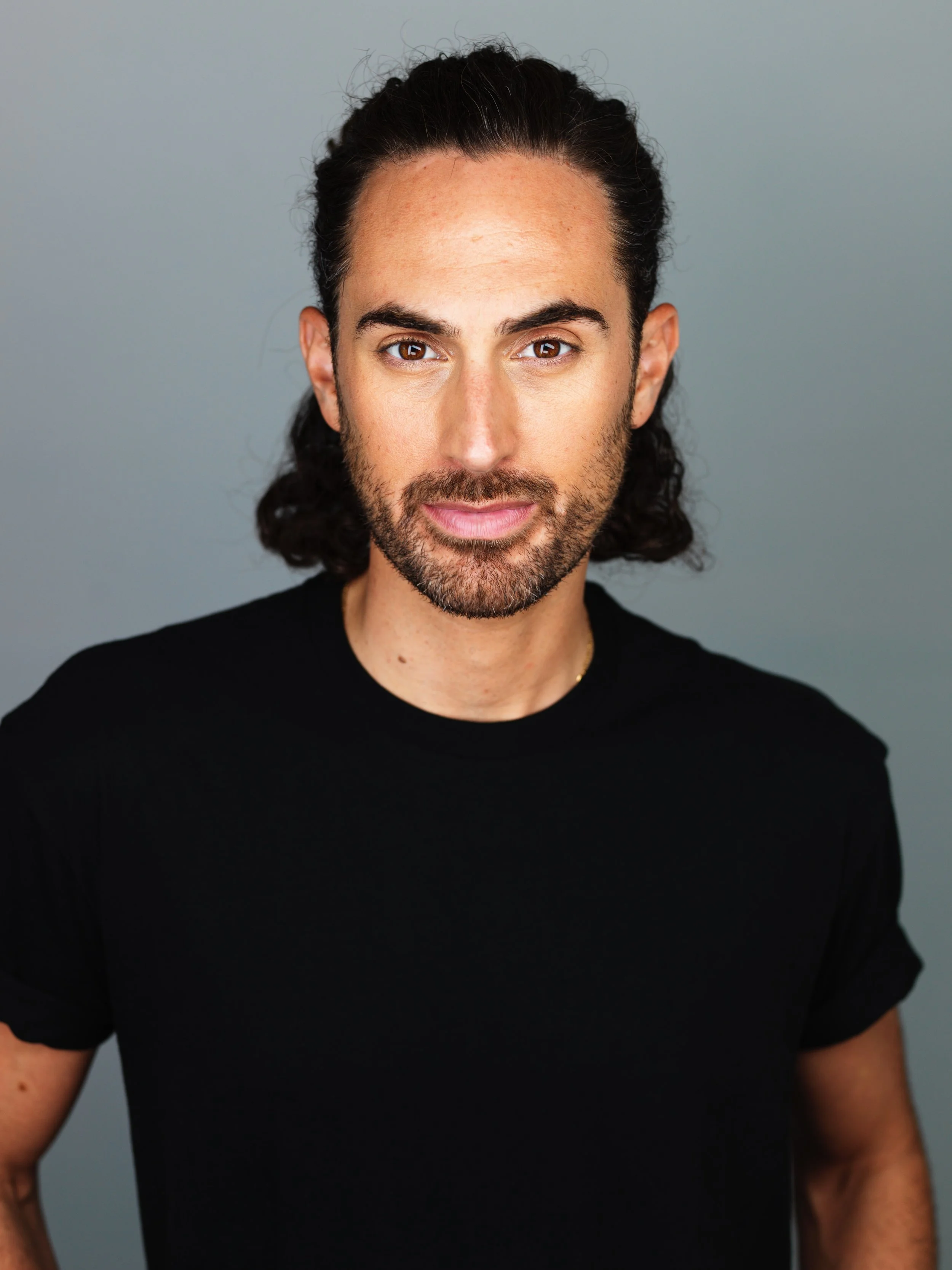 A portrait of a man with long dark hair, a beard, wearing a black t-shirt, standing against a gray background.