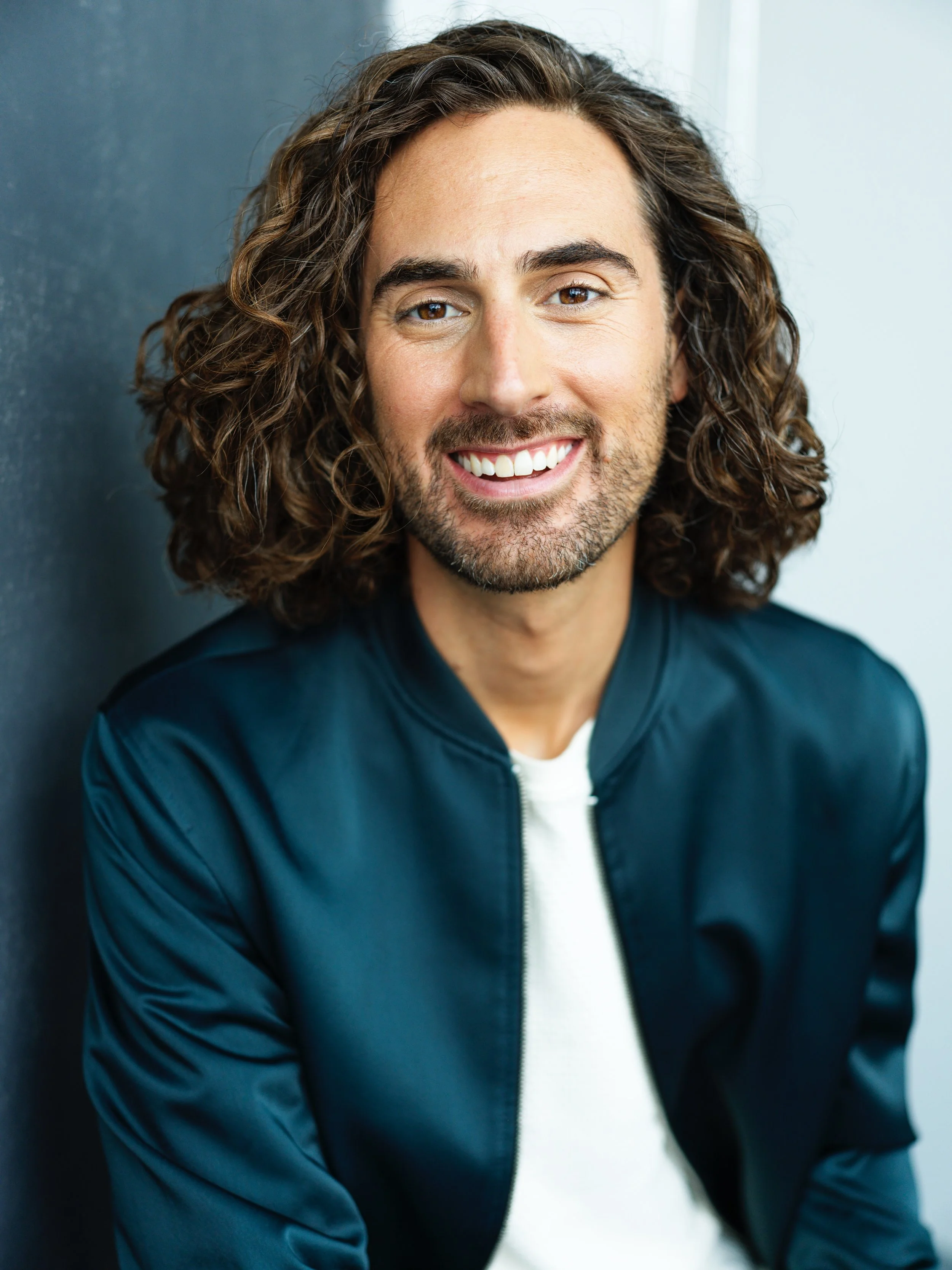 A man with curly brown hair and a beard, smiling, wearing a navy blue jacket and a white shirt, leaning against a painted wall.