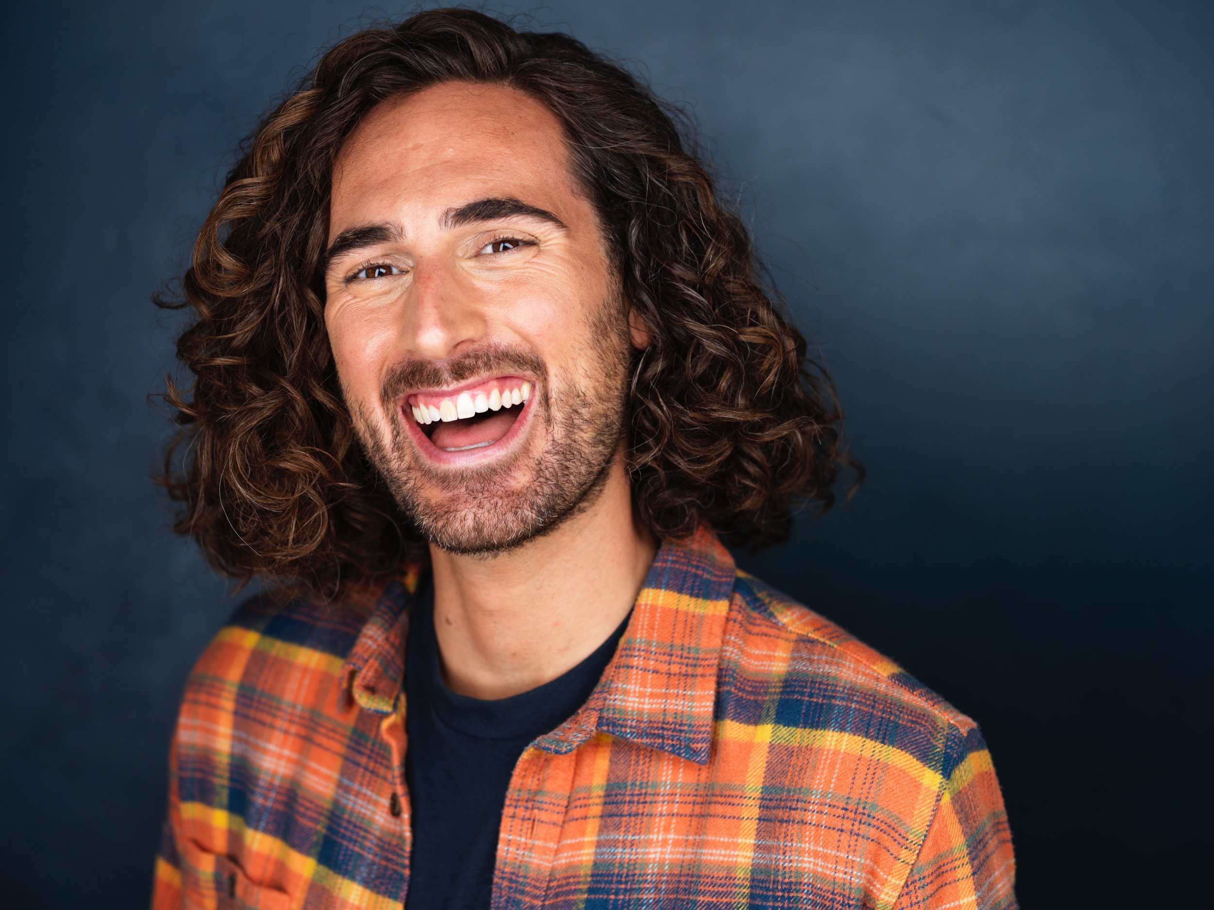 A man with medium-length curly hair and a beard, smiling widely, wearing a plaid shirt over a dark t-shirt, against a dark background.