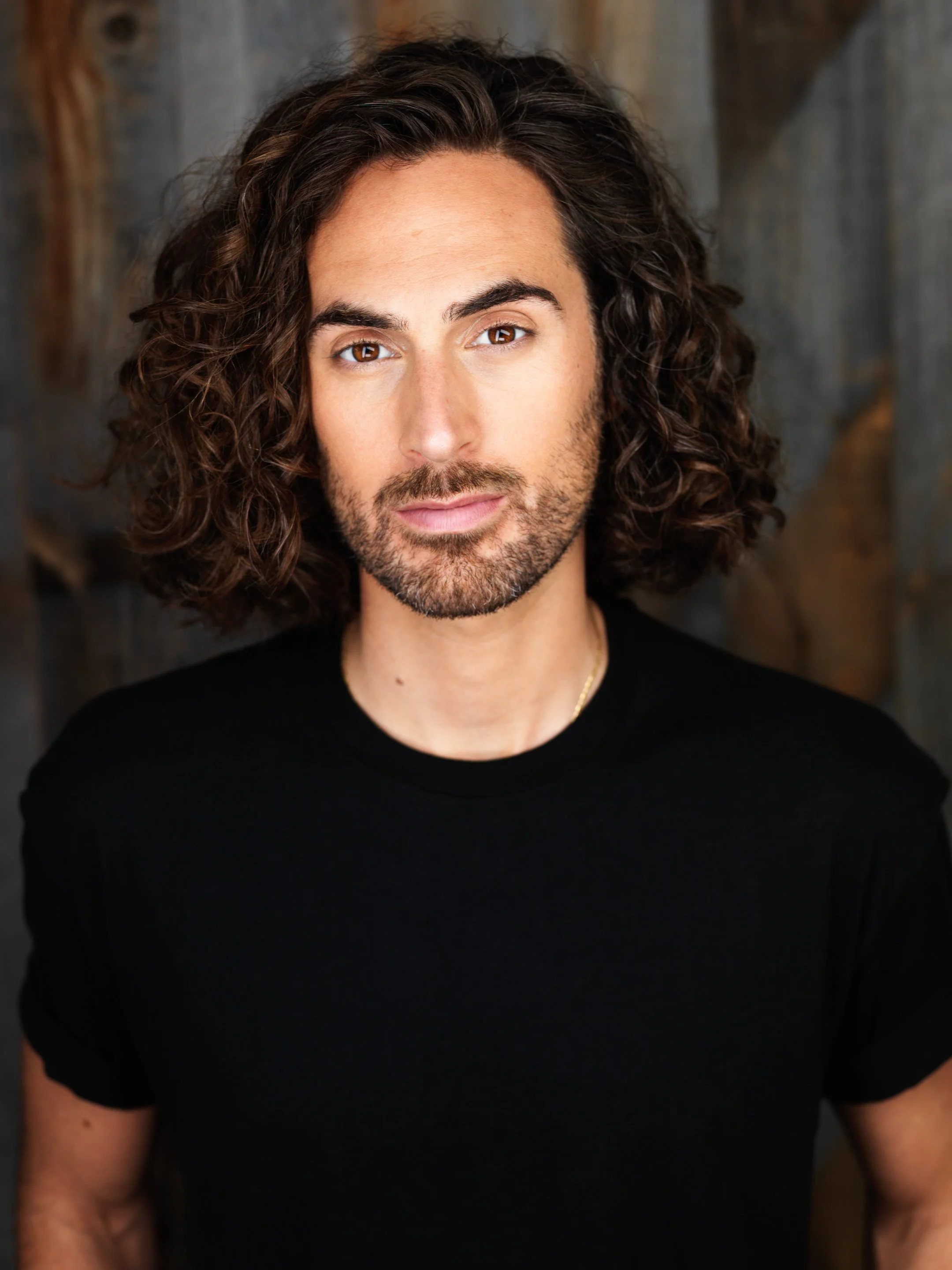 A man with long curly brown hair, a beard, and brown eyes wearing a black t-shirt, standing in front of a wooden background.