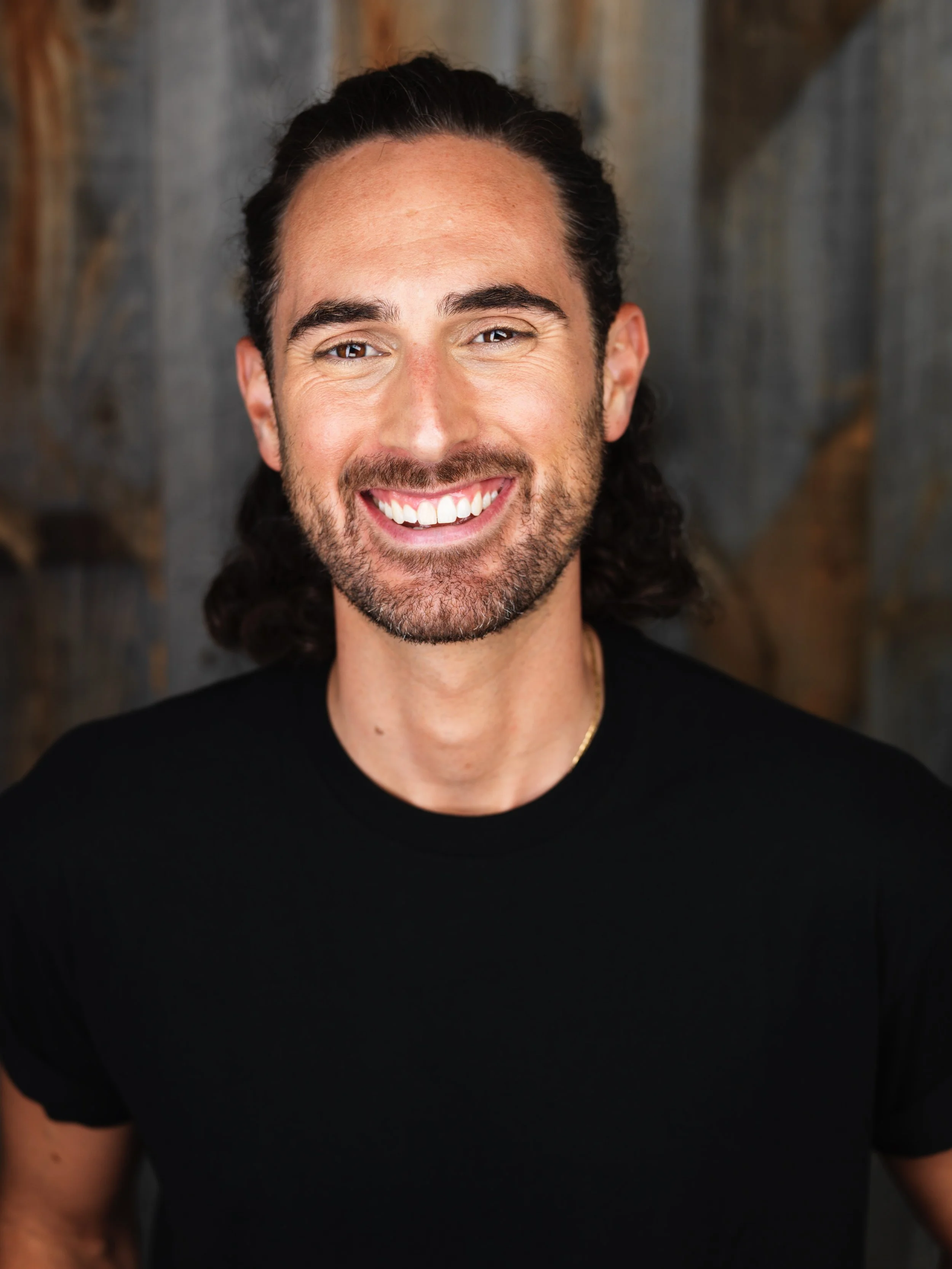 A smiling man with dark, shoulder-length curly hair, a beard, wearing a black t-shirt, standing in front of a wooden background.