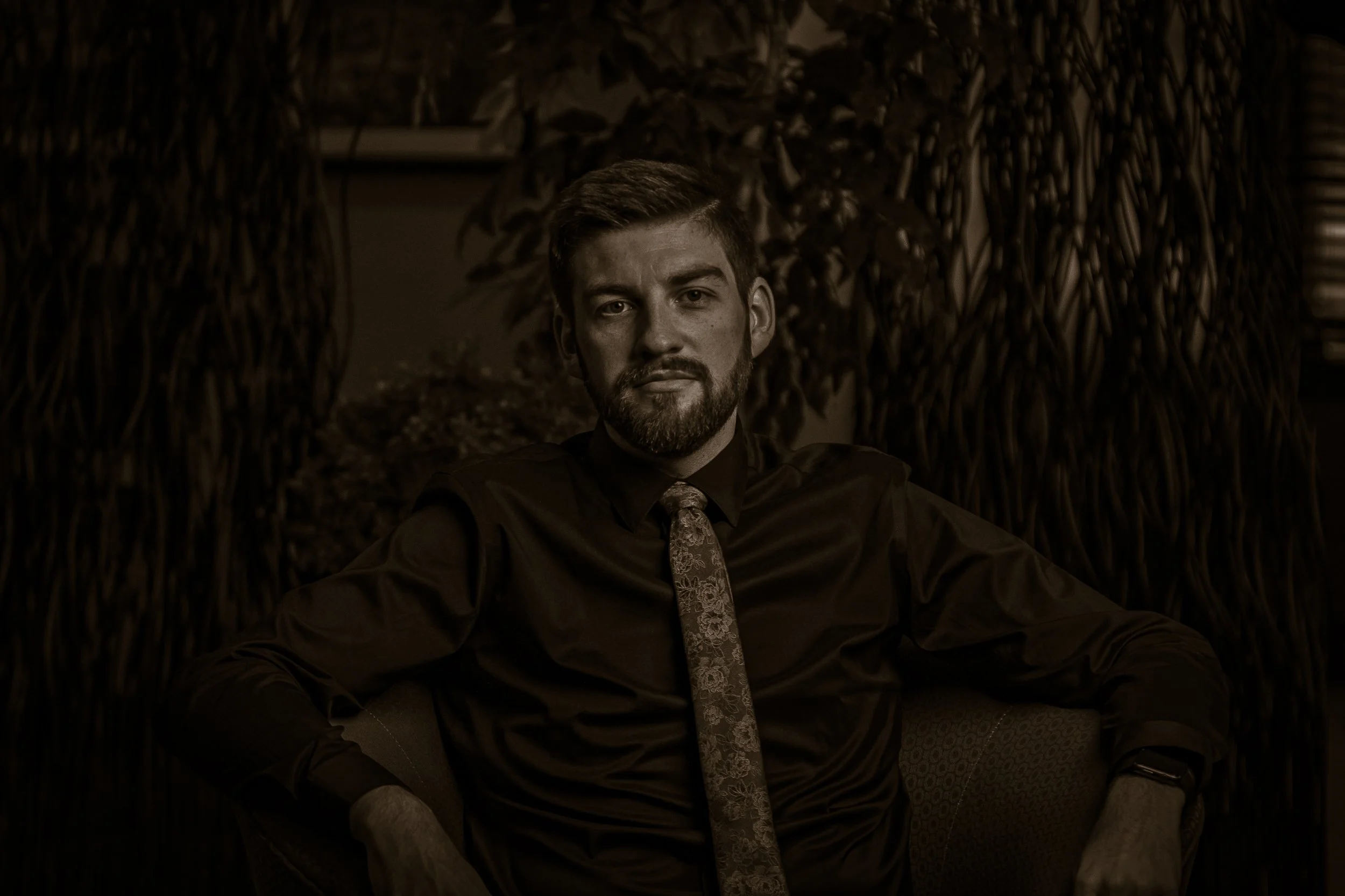 A man with a beard, wearing a dark shirt and patterned tie, sitting in front of a leafy background.