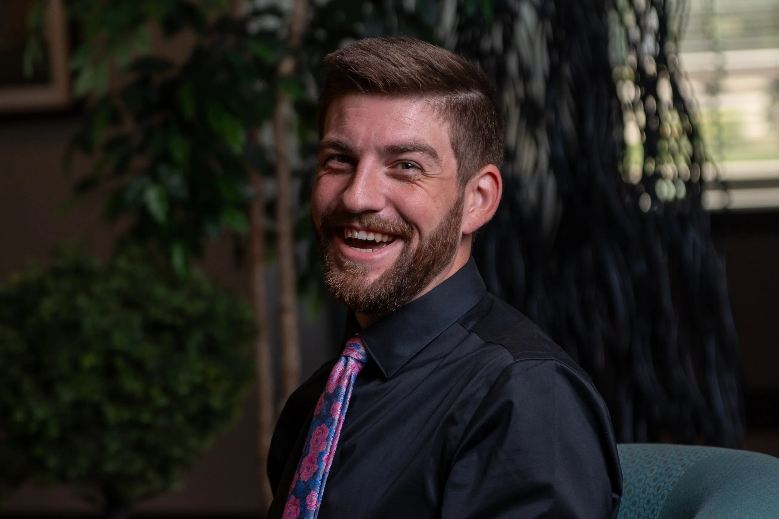 Man with short brown hair and beard smiling, wearing a dark shirt and colorful tie, sitting indoors with plants and a window with blinds