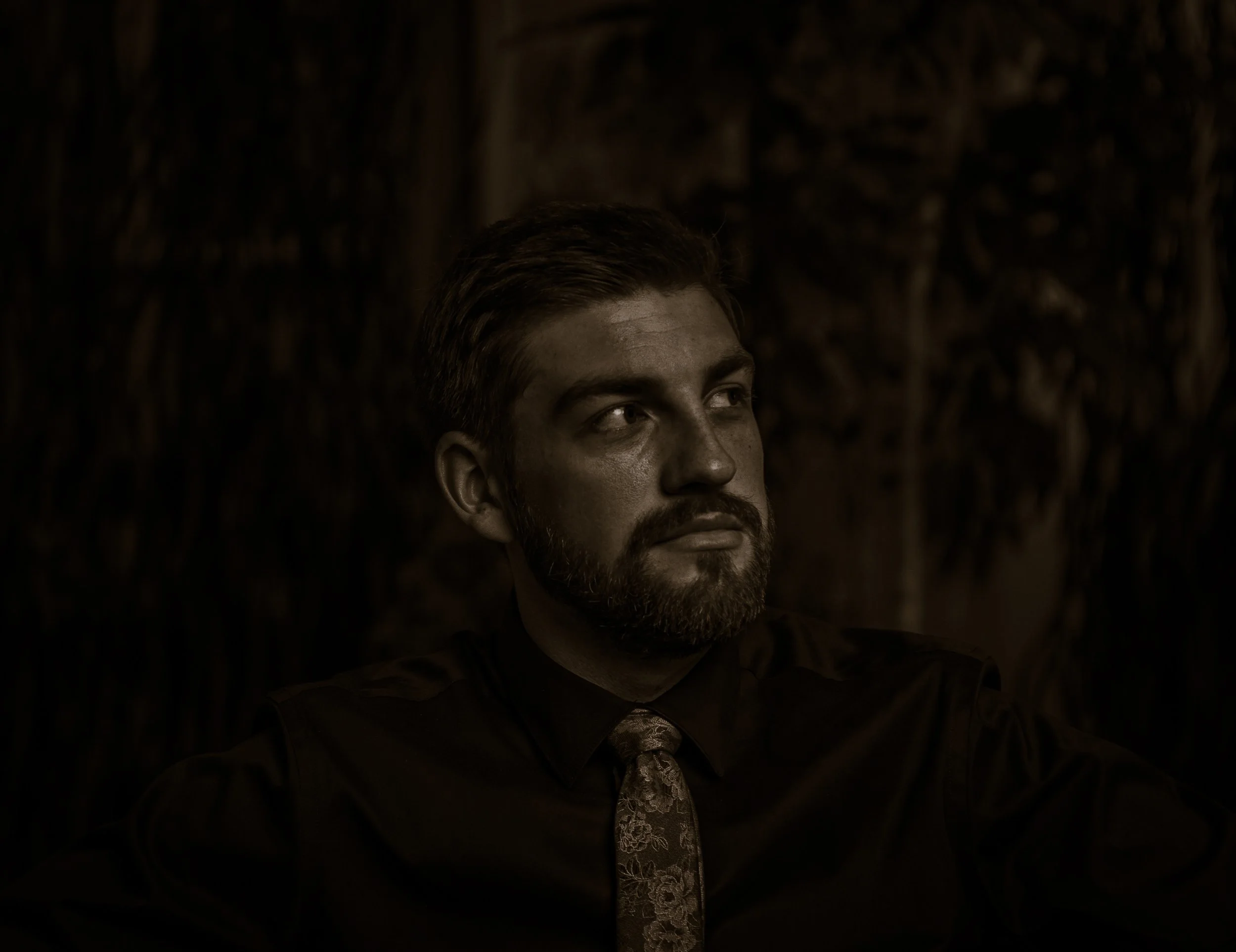 A man with a beard and slicked-back hair wearing a dark shirt and patterned tie, looking to the side, against a dark textured background.