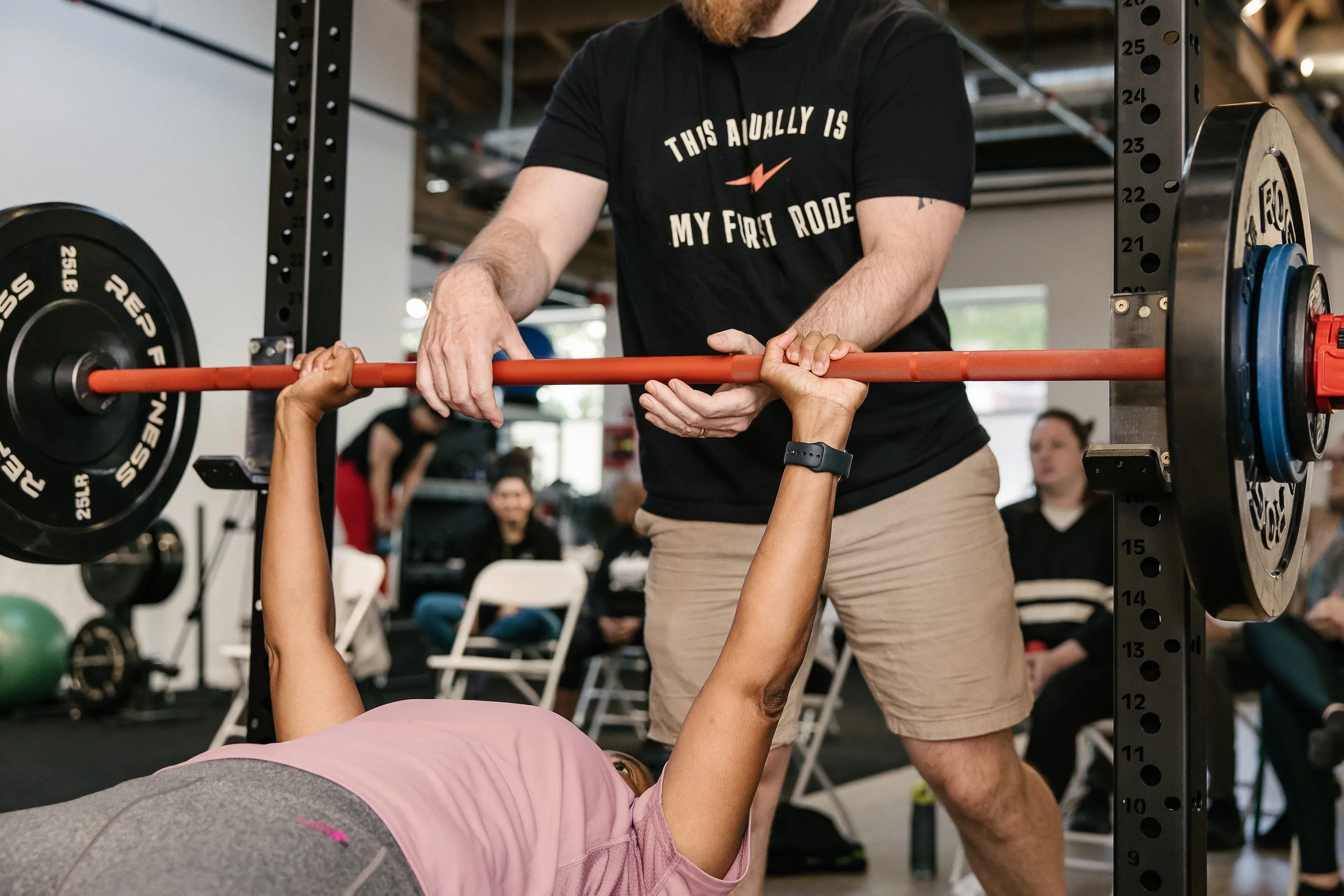 A female lifter wearing a pink shirt lays on a bench pressing the barbell.