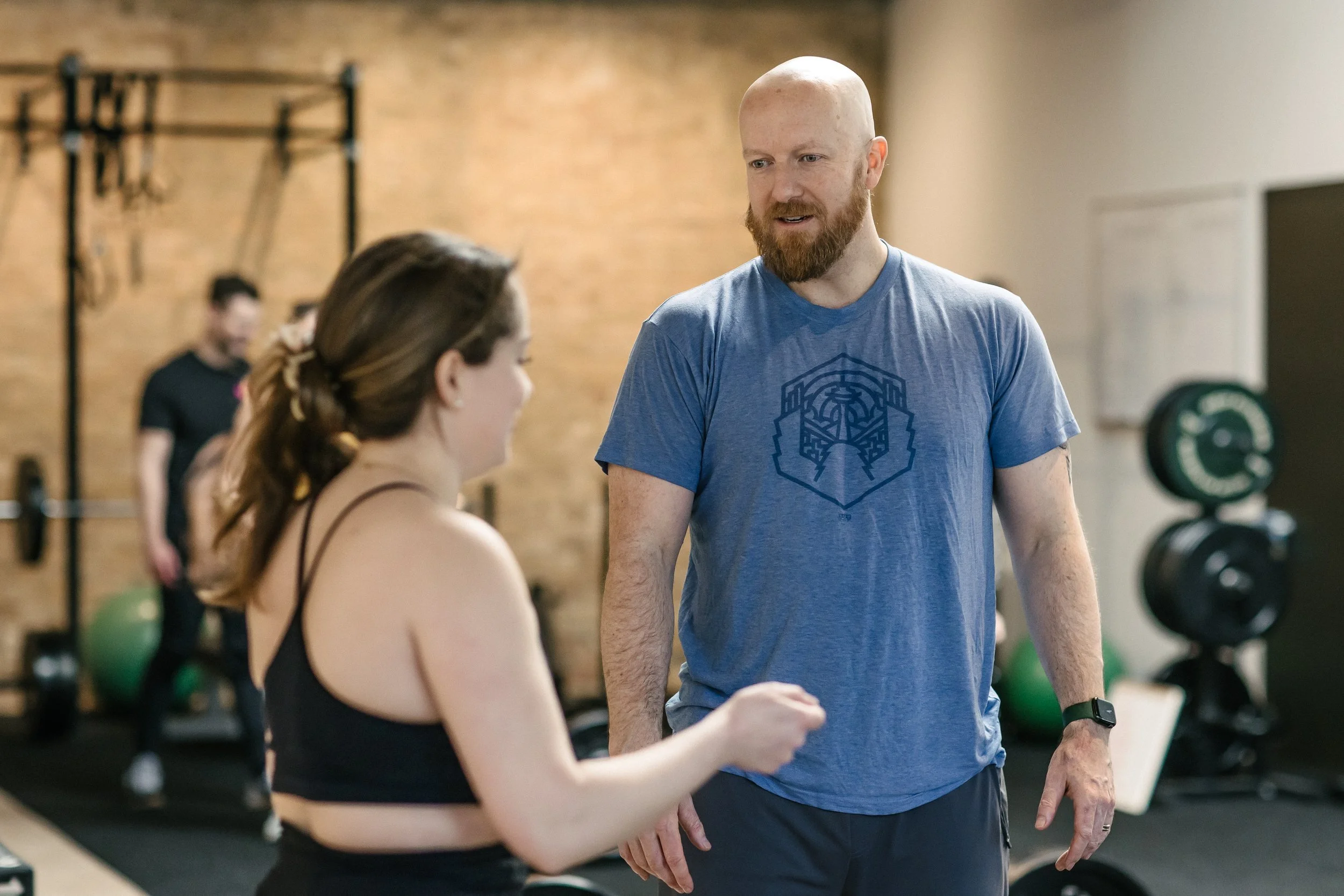 A man with a beard and a shaved head, wearing a blue t-shirt, is talking to a woman with long brown hair tied back, wearing a black sports bra, in a gym setting.