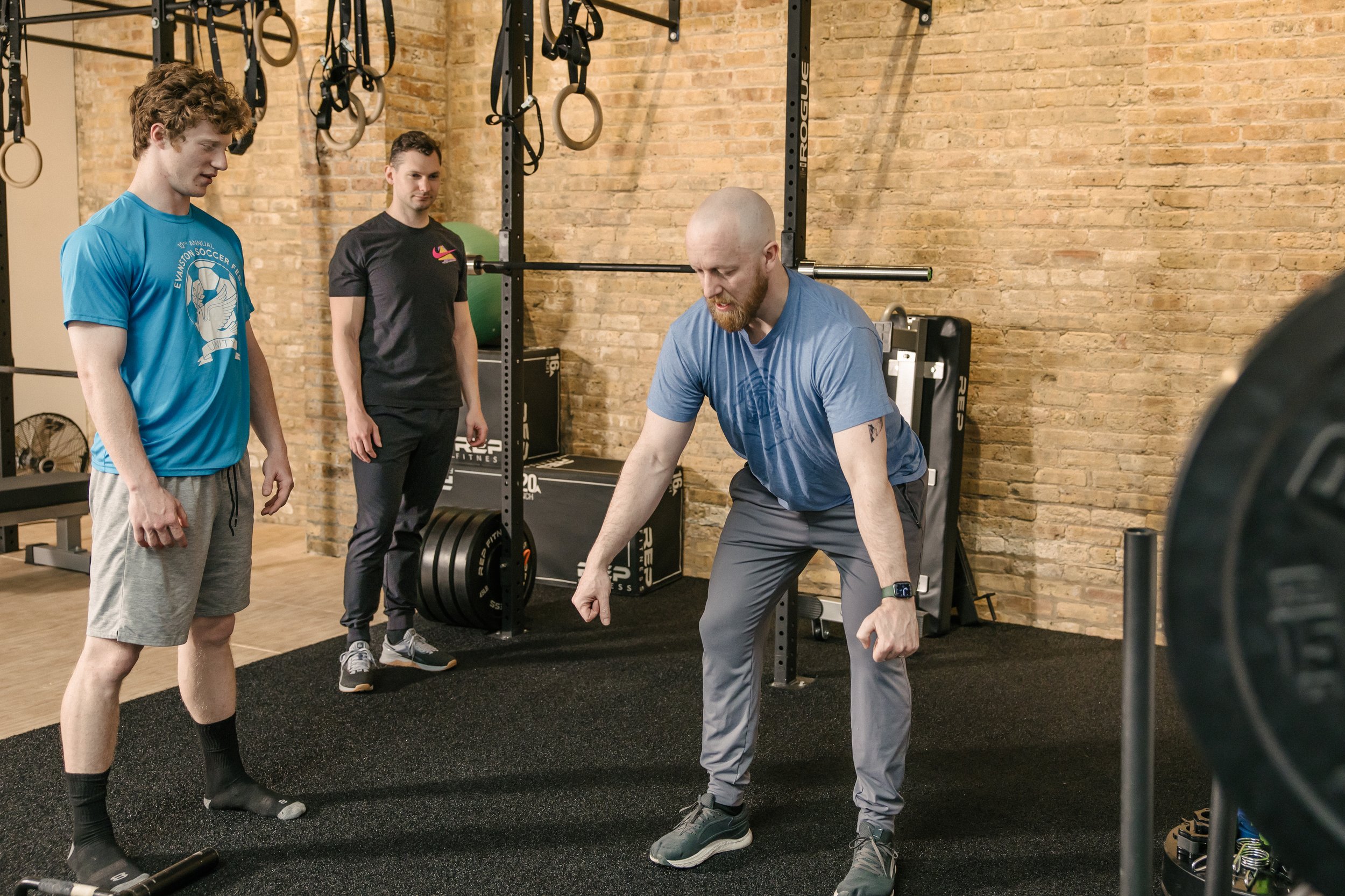 A group of three men in a gym with brick walls, one man is squatting with weight plates, and the other two are observing him.