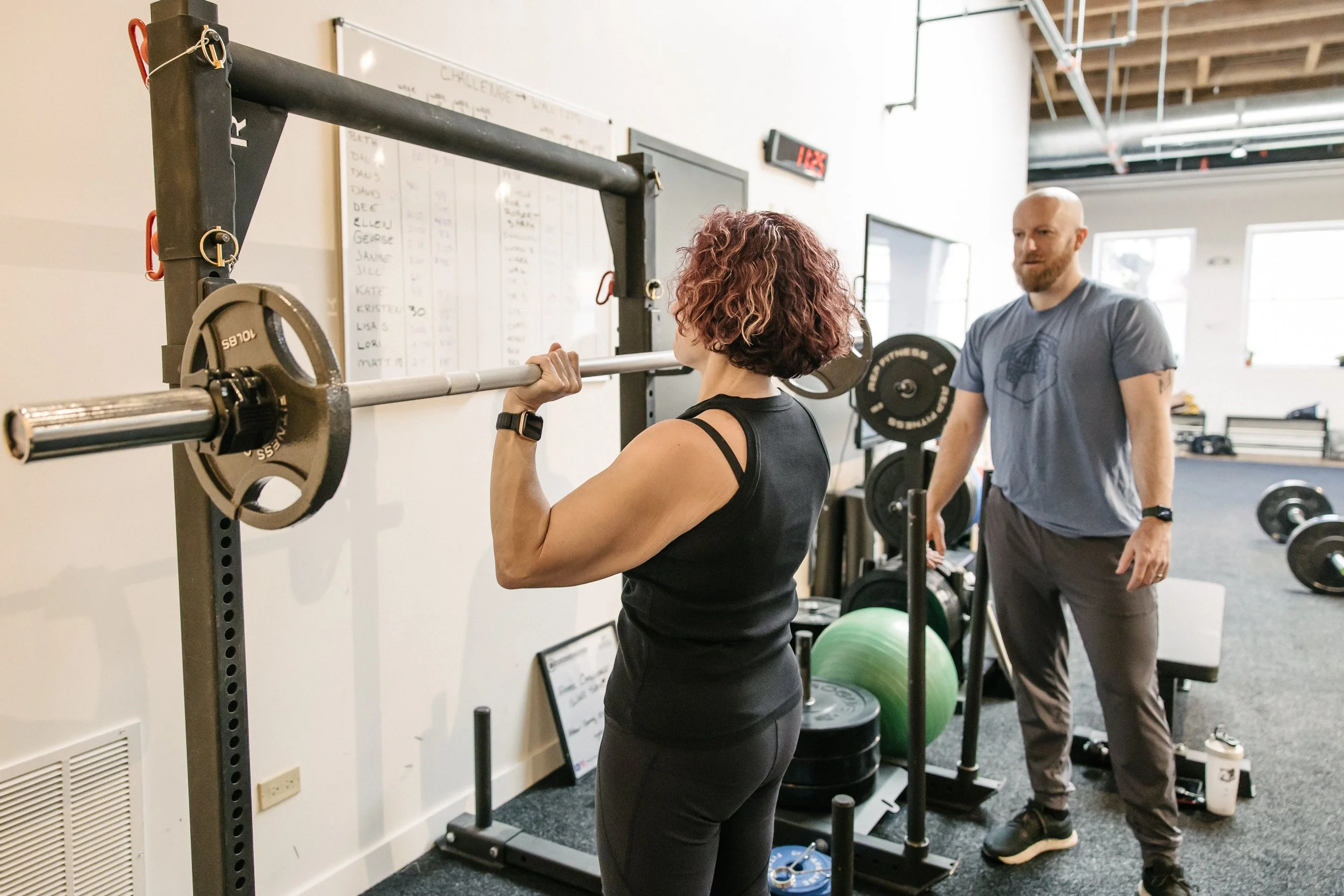 Woman with short curly hair lifting a barbell during a weightlifting session supervised by trainer in a gym.