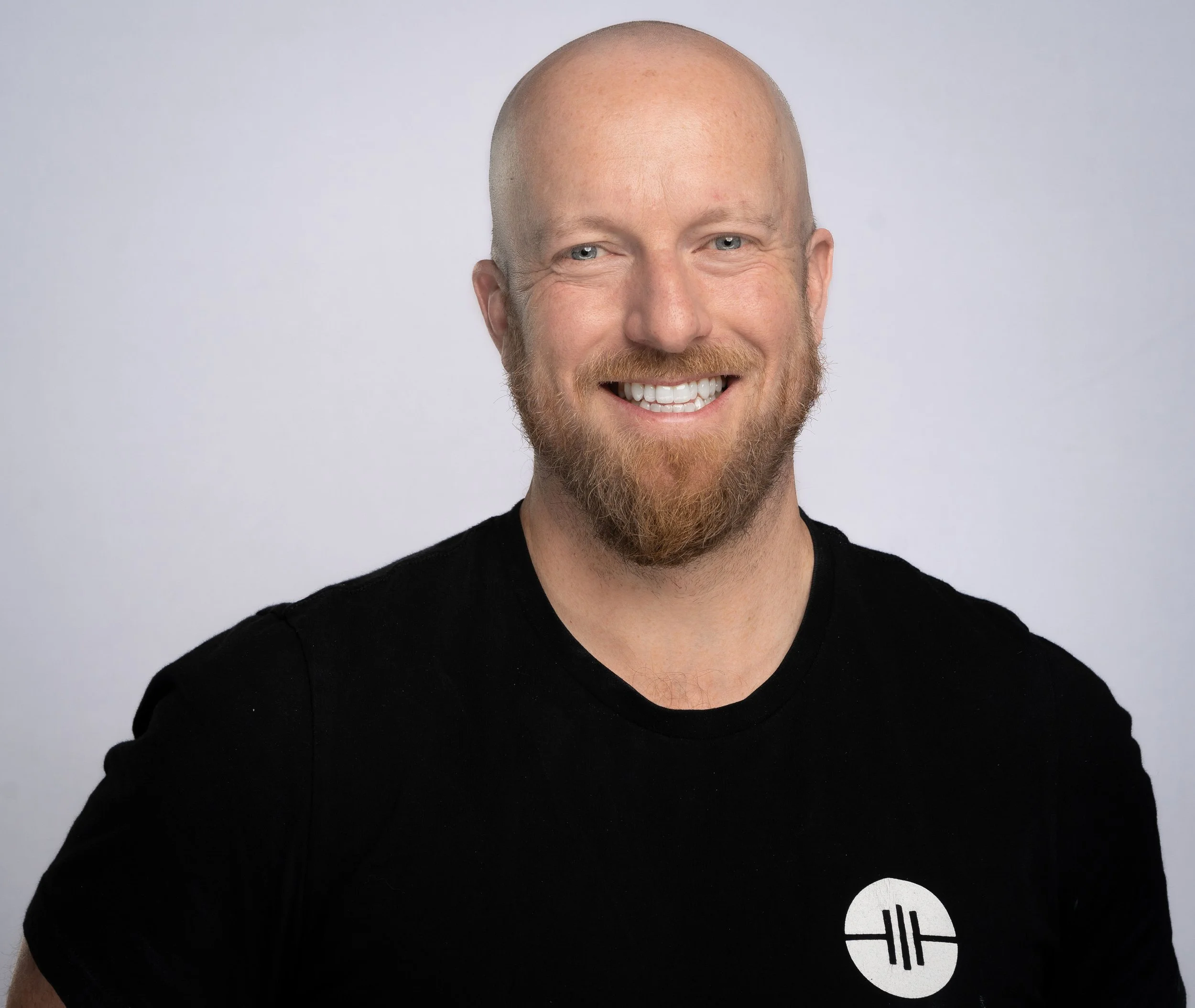 A smiling man with a beard and mustache, wearing a black T-shirt with a logo on the chest, against a plain light gray background.