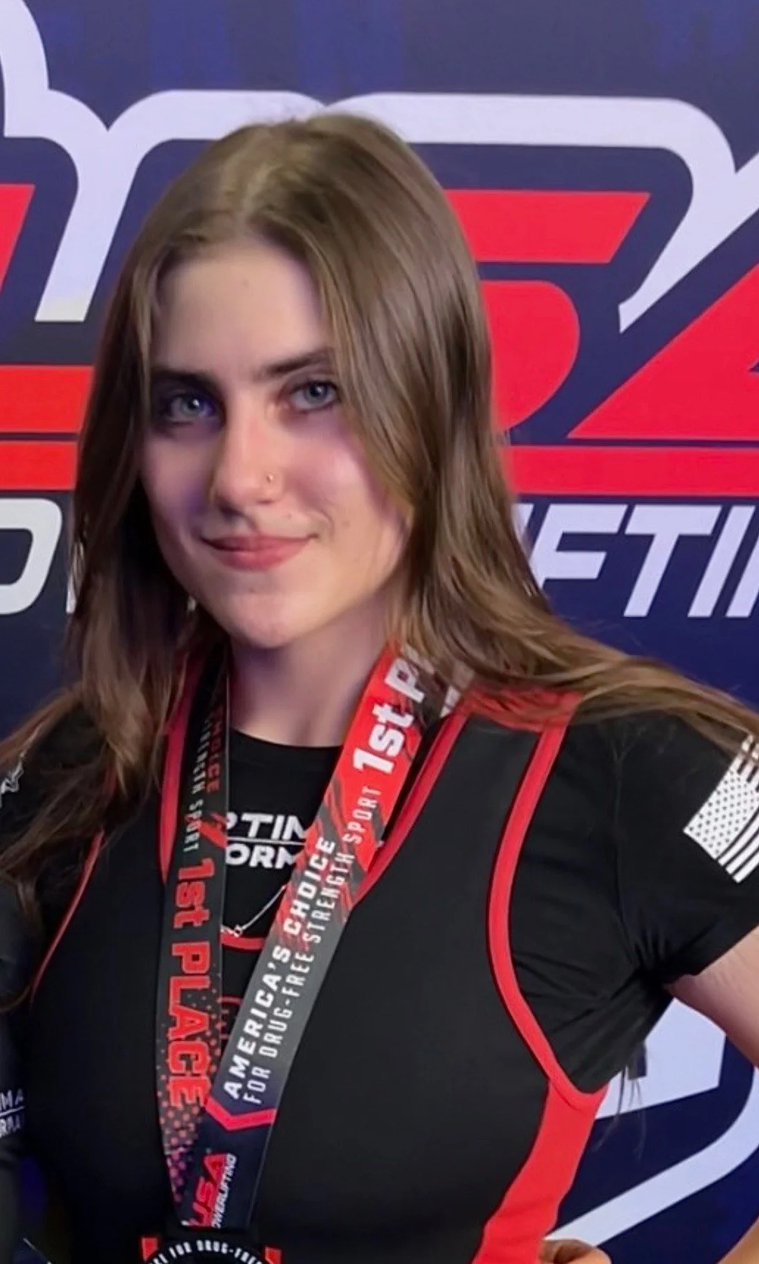 A young woman with long brown hair and a nose piercing, wearing a black and red racing shirt and a medal around her neck, standing in front of a blue and red racing-themed backdrop.