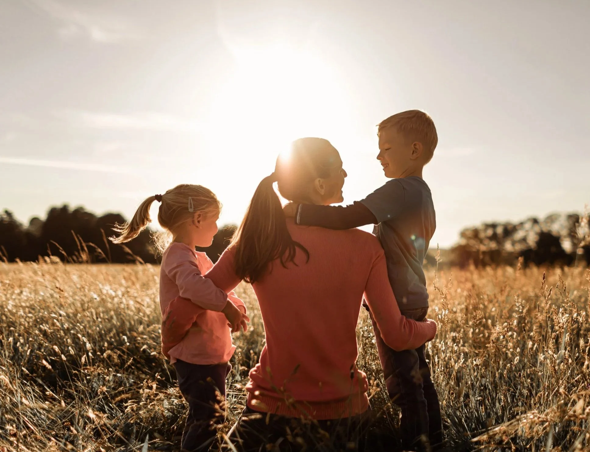 A woman and three children in a field during sunset, with the woman holding a boy in her arms and a girl standing nearby.