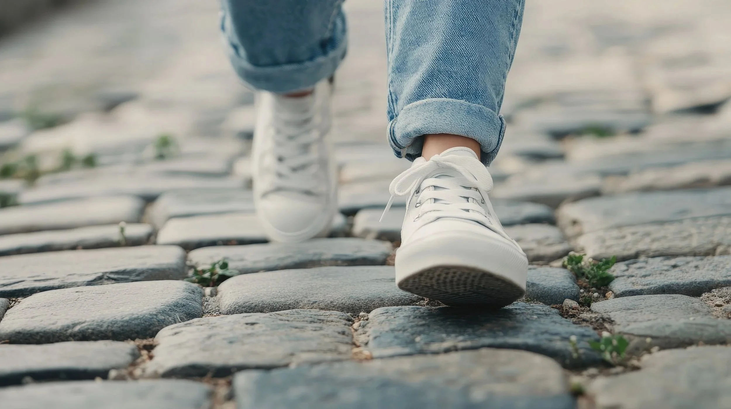 Close-up of someone walking on cobblestone street wearing white sneakers and blue rolled-up jeans.
