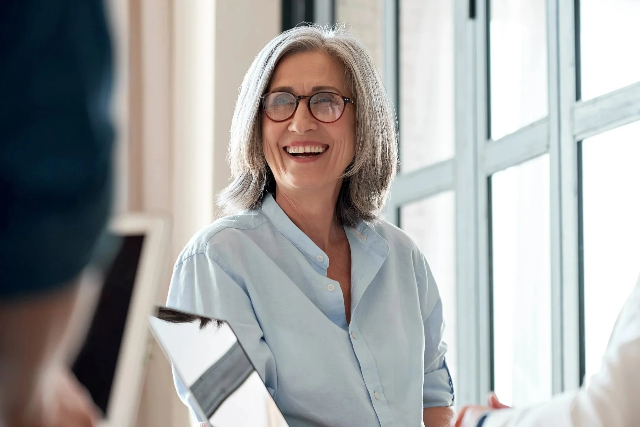 A smiling older woman with gray hair and glasses, wearing a light blue shirt, engaged in a conversation with someone off-camera near large windows.