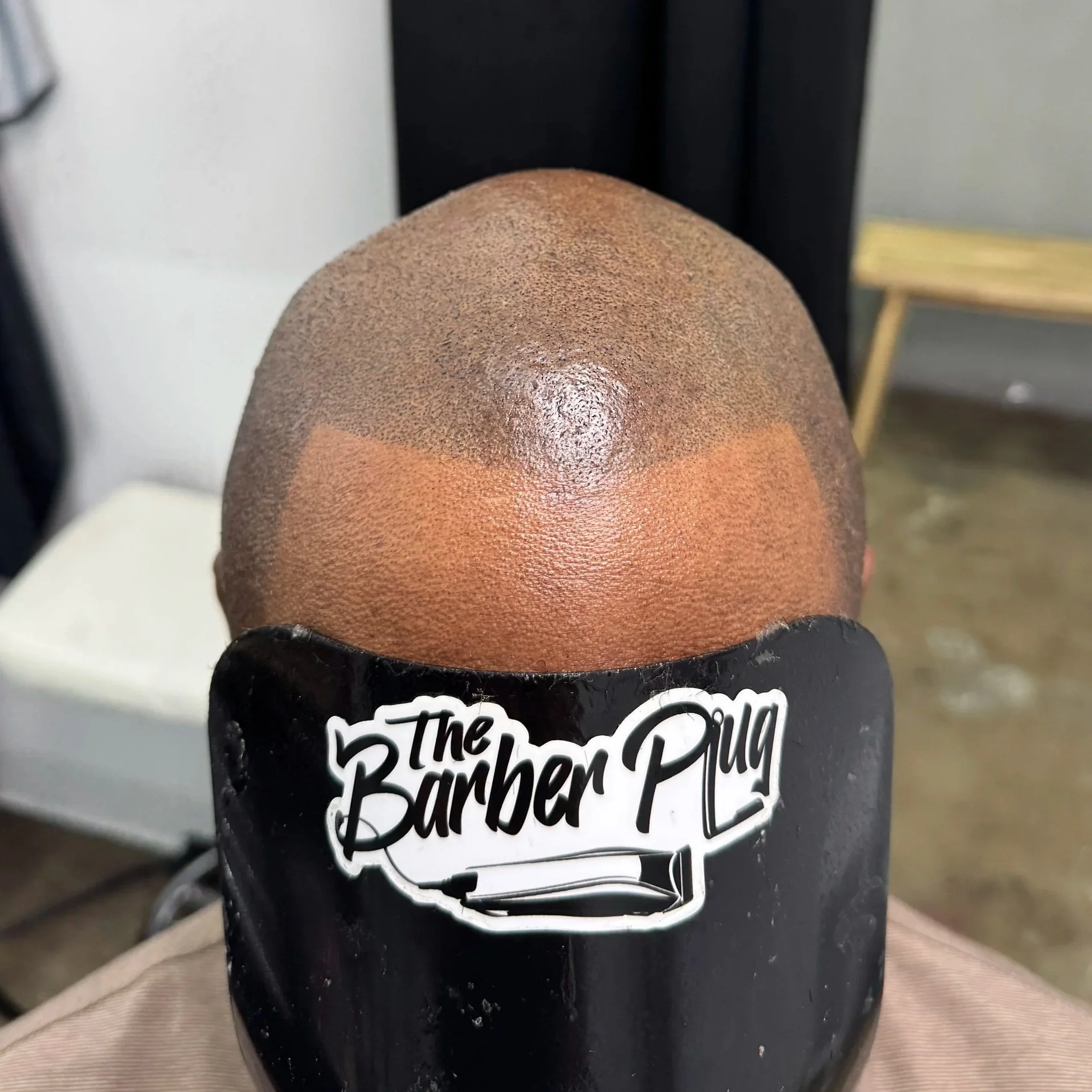 Close-up of a person's head with freshly shaved, textured hair and a fade haircut, sitting in a barbershop chair with a black headrest that has a sticker reading 'The Barber Plug'.