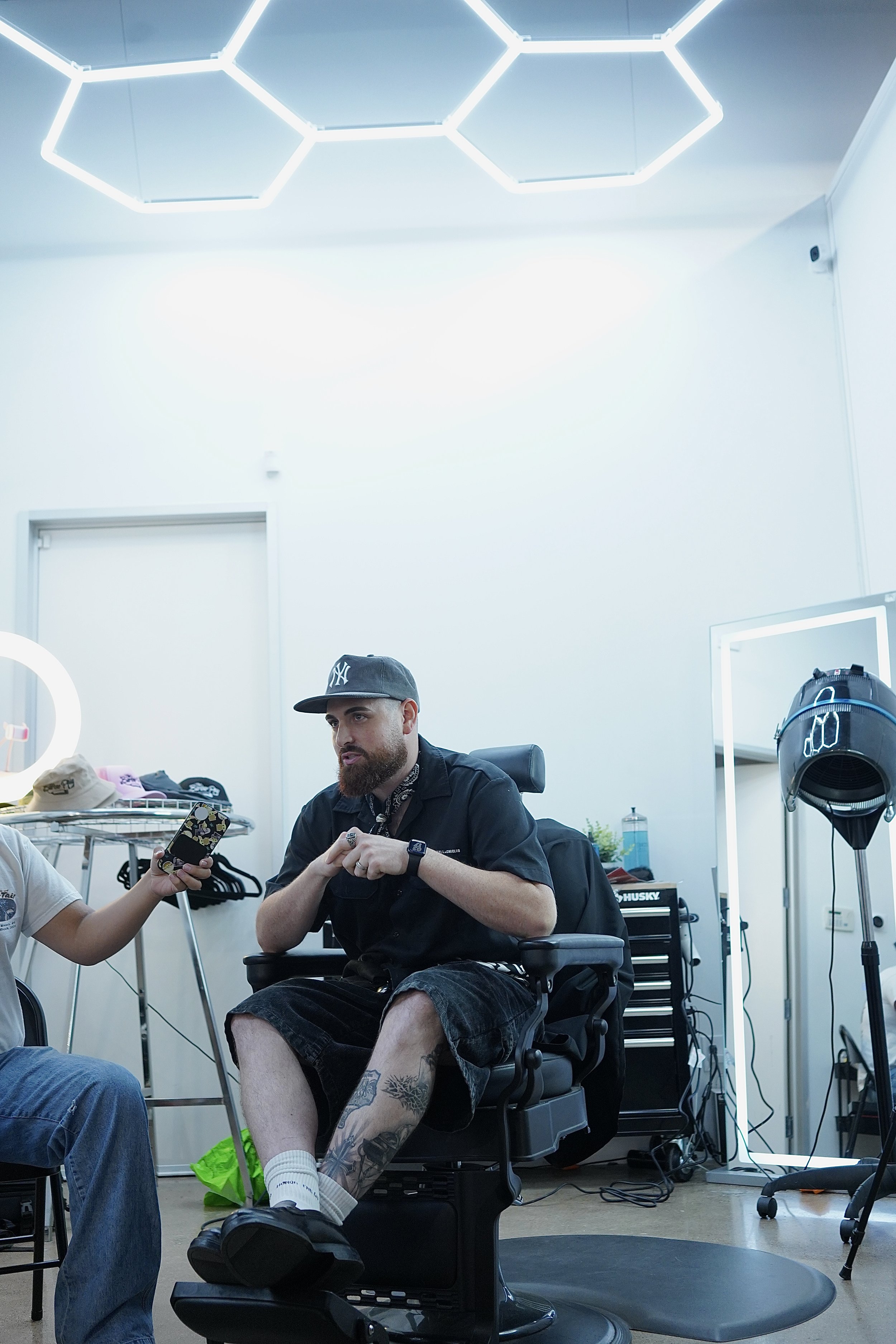 A man with a beard, wearing a baseball cap, black shirt, shorts, and white socks, sitting in a black barber chair in a brightly lit salon or studio with modern lighting, a mirror, and hairstyling tools visible.