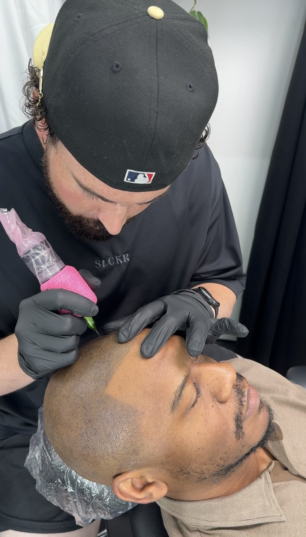 A tattoo artist is performing a tattoo on a man's forehead, who is lying down with his eyes closed. The artist is wearing black gloves and a black cap with the MLB logo, and is holding a tattoo machine with a pink cover.
