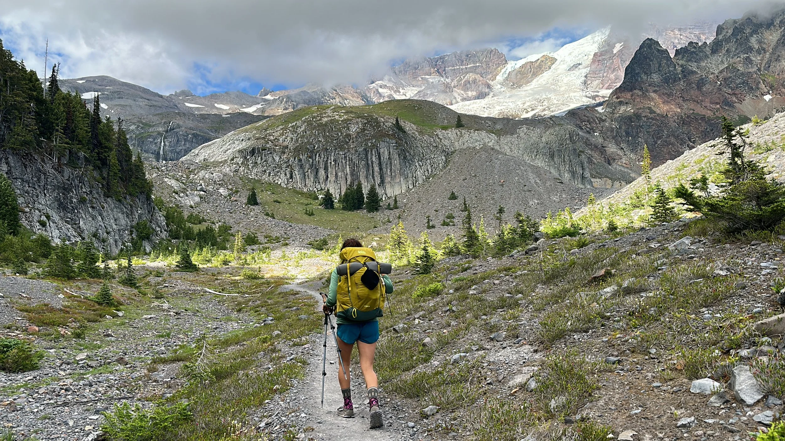 A person hiking with a backpack and trekking poles on a rocky trail in a mountainous landscape with trees, rocky terrain, and snow-capped peaks in the background.