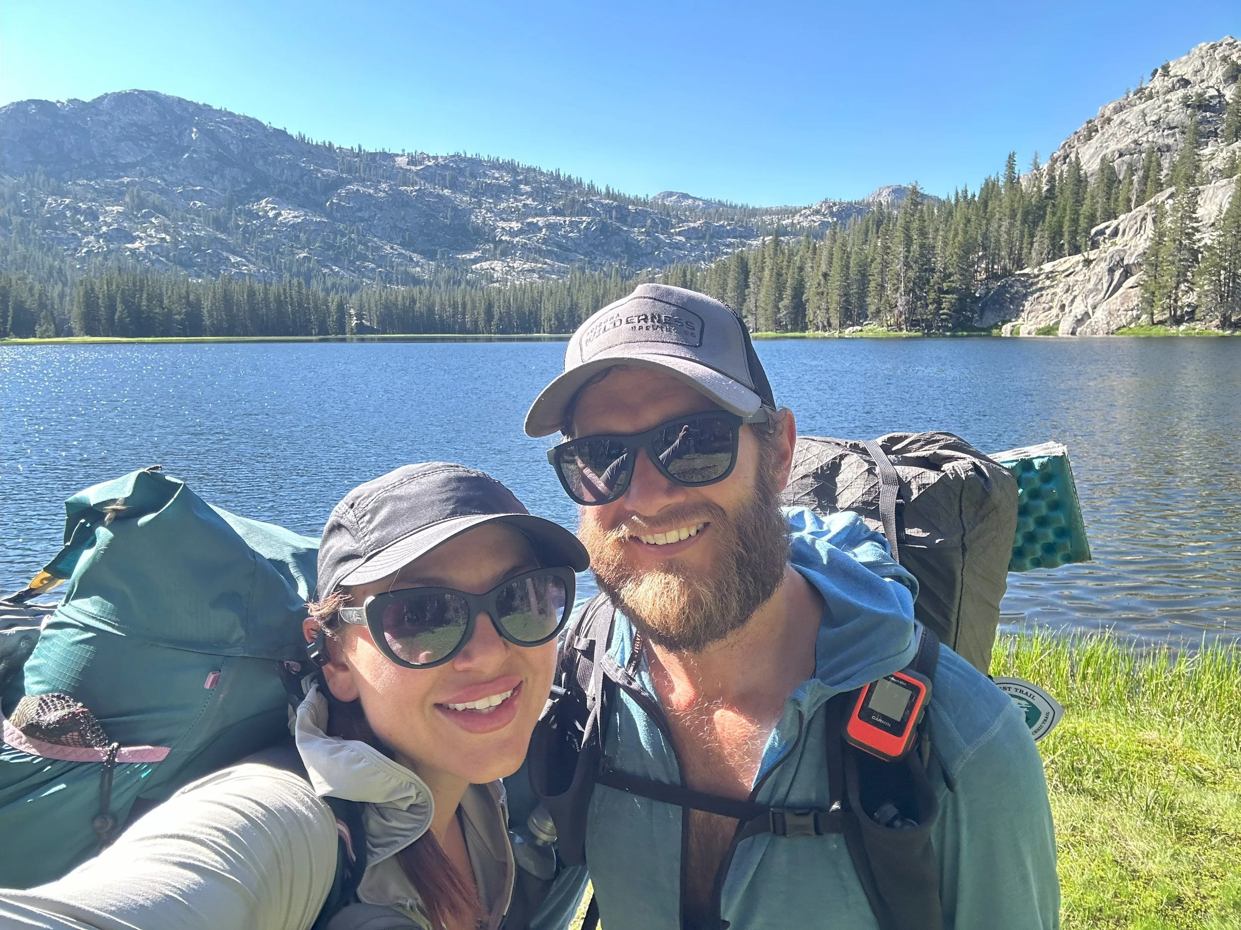 A smiling couple taking a selfie outdoors near a lake with mountains and trees in the background on a bright, sunny day.