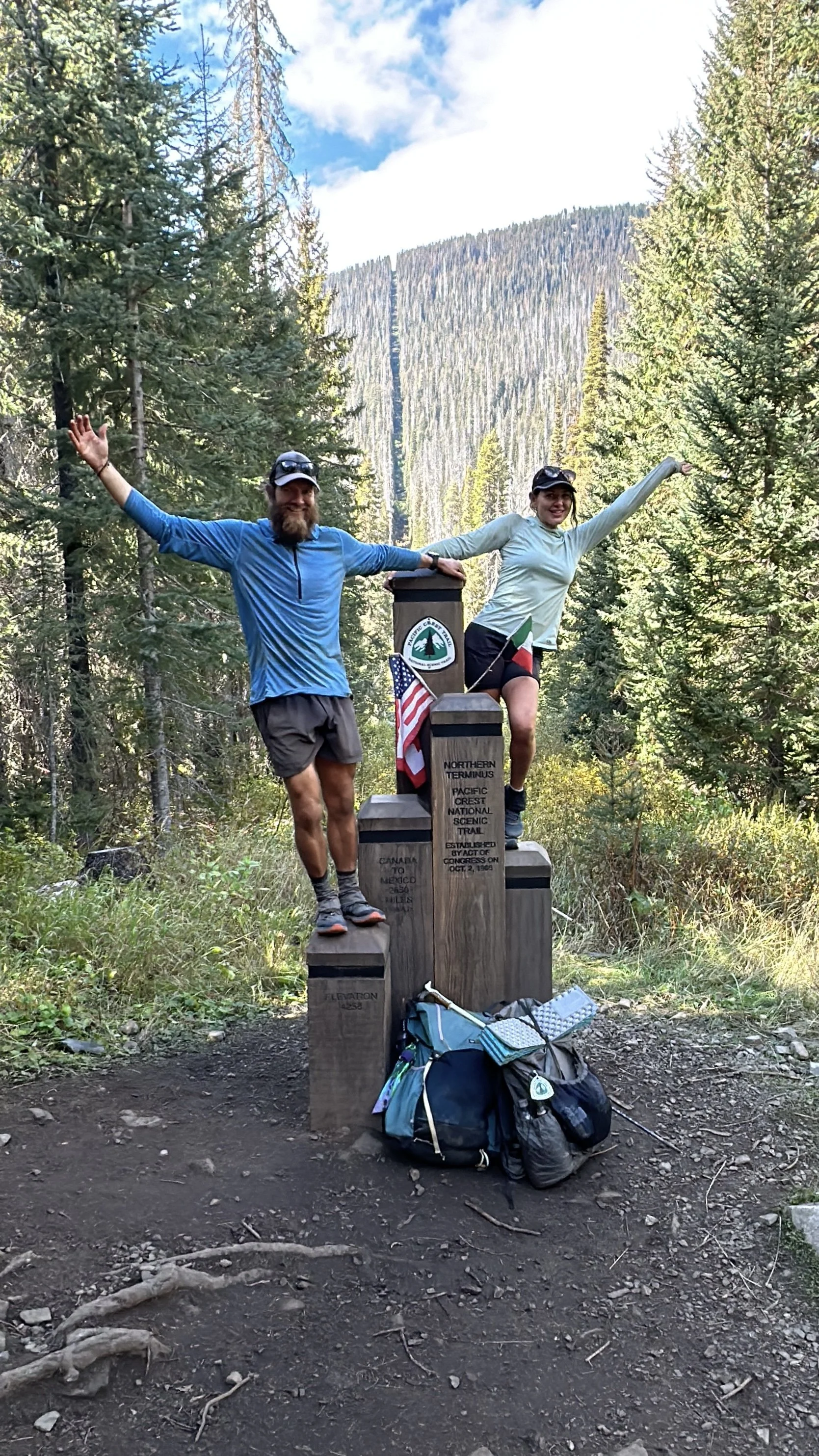 Two hikers standing on the Northern Terminus of the Pacific Crest National Scenic Trail, holding hands and smiling with a backpack on the ground in front. The trail marker indicates the end of the trail, with trees and mountains in the background.