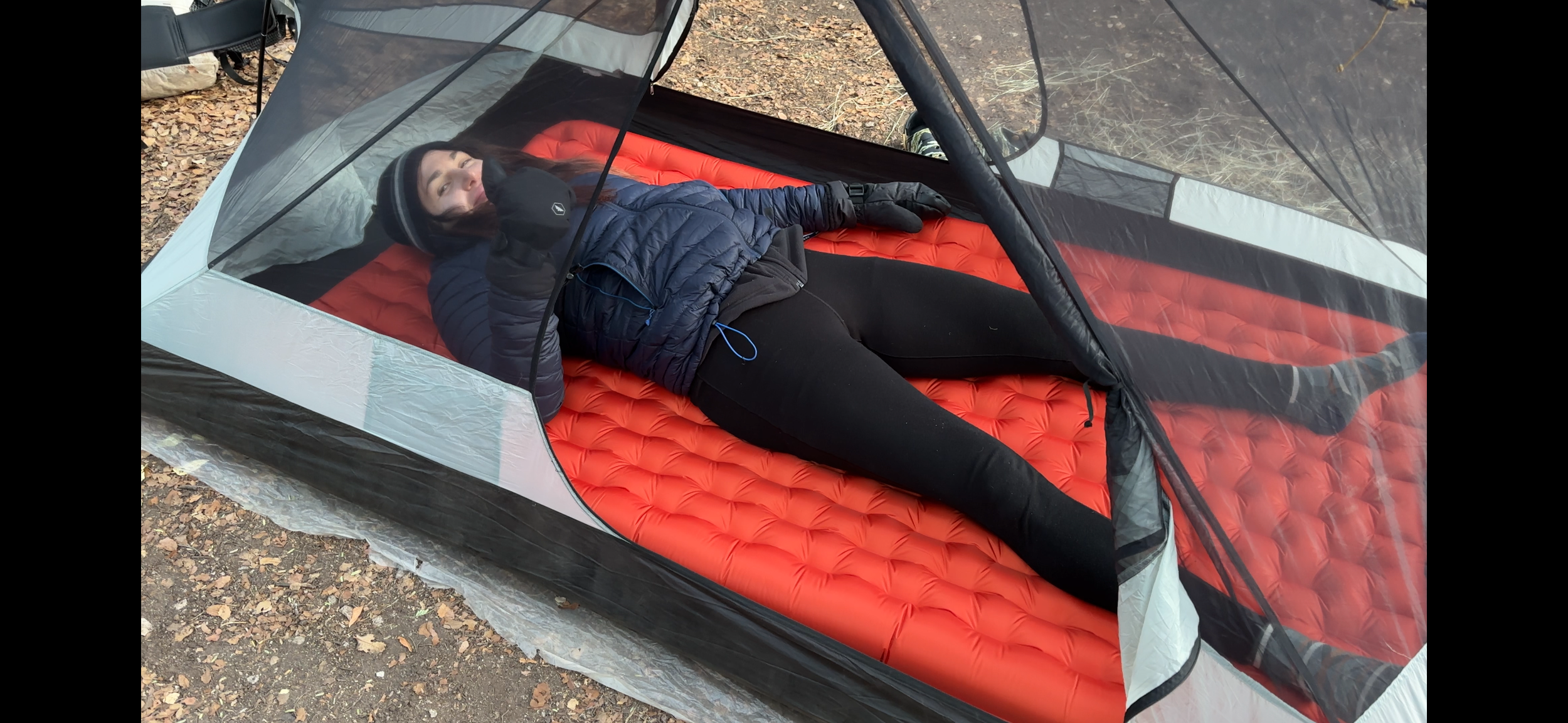Person lying inside a tent on a red sleeping pad, bundled in warm clothing, giving a thumbs up.