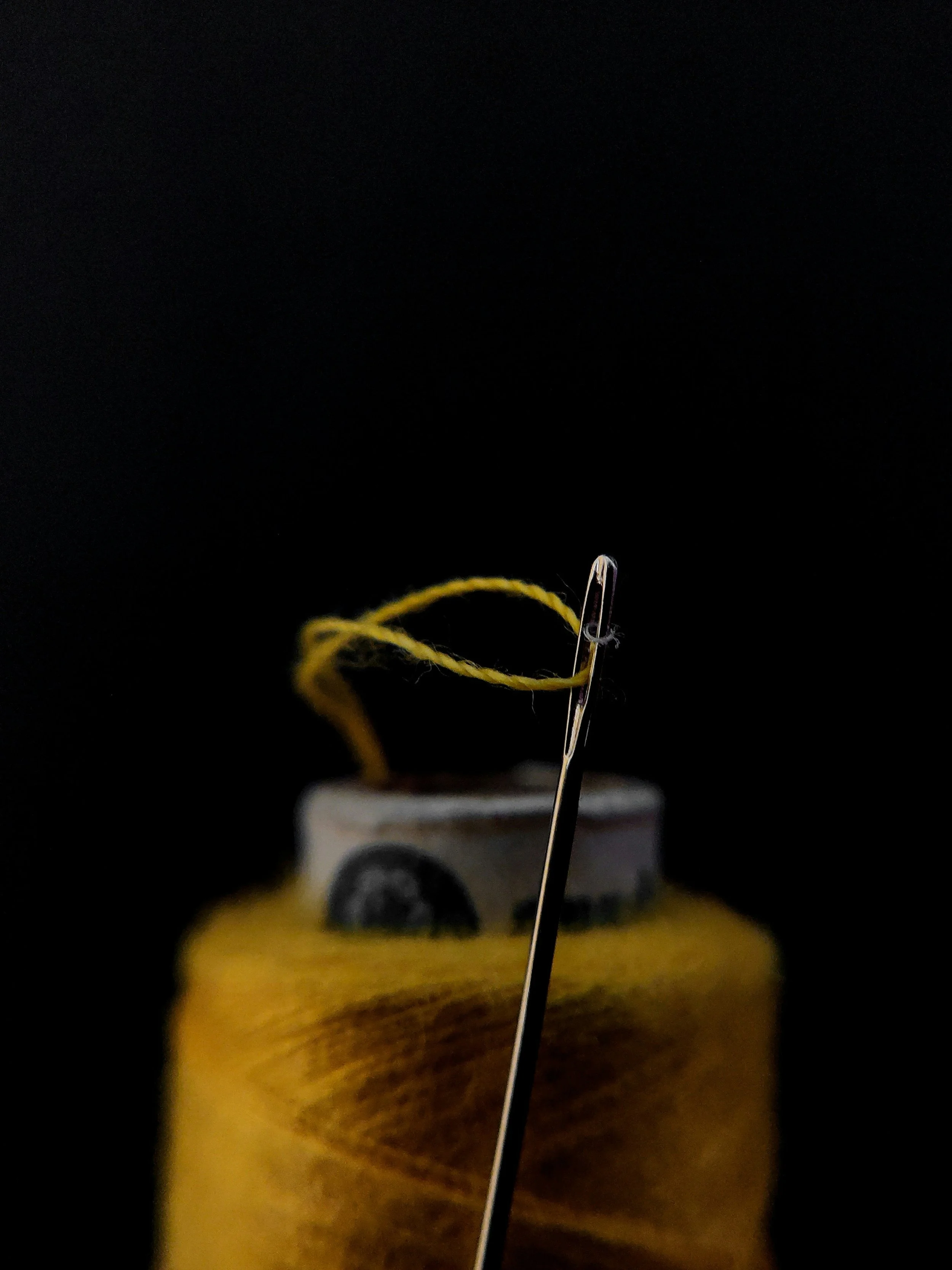 Close-up of a sewing needle with yellow thread passing through its eye, inserted into a wooden spool of yellow thread against a black background.