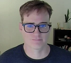 A person with light brown hair, wearing glasses and a dark shirt, poses in front of a light-colored wall with a small plant and a bookshelf in the background.