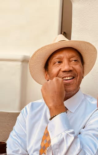 Man wearing a light blue shirt, orange checkered tie, and beige hat, smiling and resting his chin on his hand.