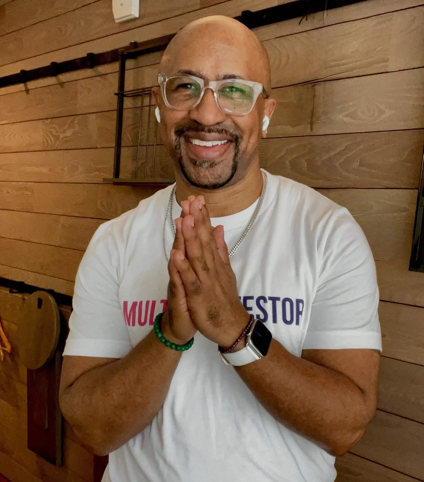 A smiling man with glasses and a goatee, wearing a white T-shirt and smartwatch, standing indoors with wooden wall paneling, clasping hands together in a gesture of gratitude or prayer.