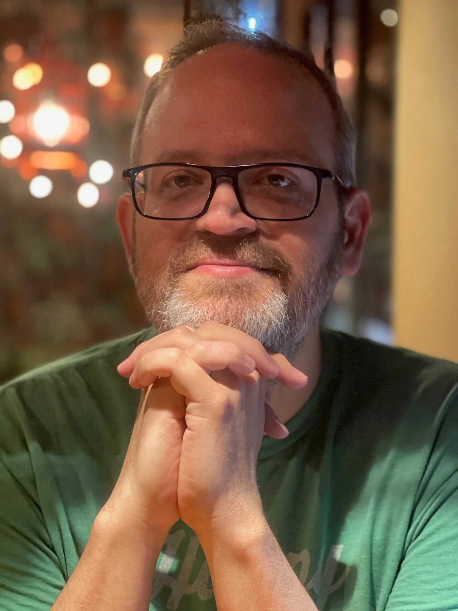 A man with glasses and a beard, sitting with hands clasped under his chin, in a warmly lit indoor setting with blurred background lights.