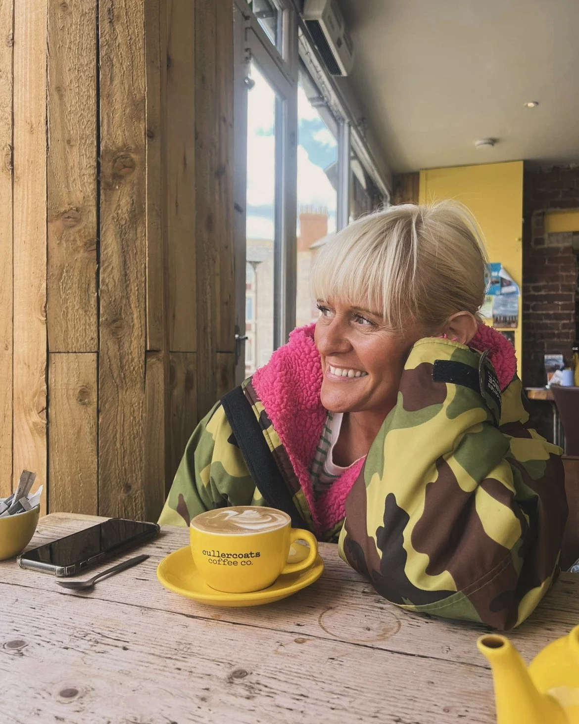 Ashley Madeliene Pigford, counsellor, is smiling and sitting at a wooden table in a cafe. She is wearing a camo jacket with a pink fleece lining. On the table in front of her, there is a yellow coffee cup with a matching saucer.