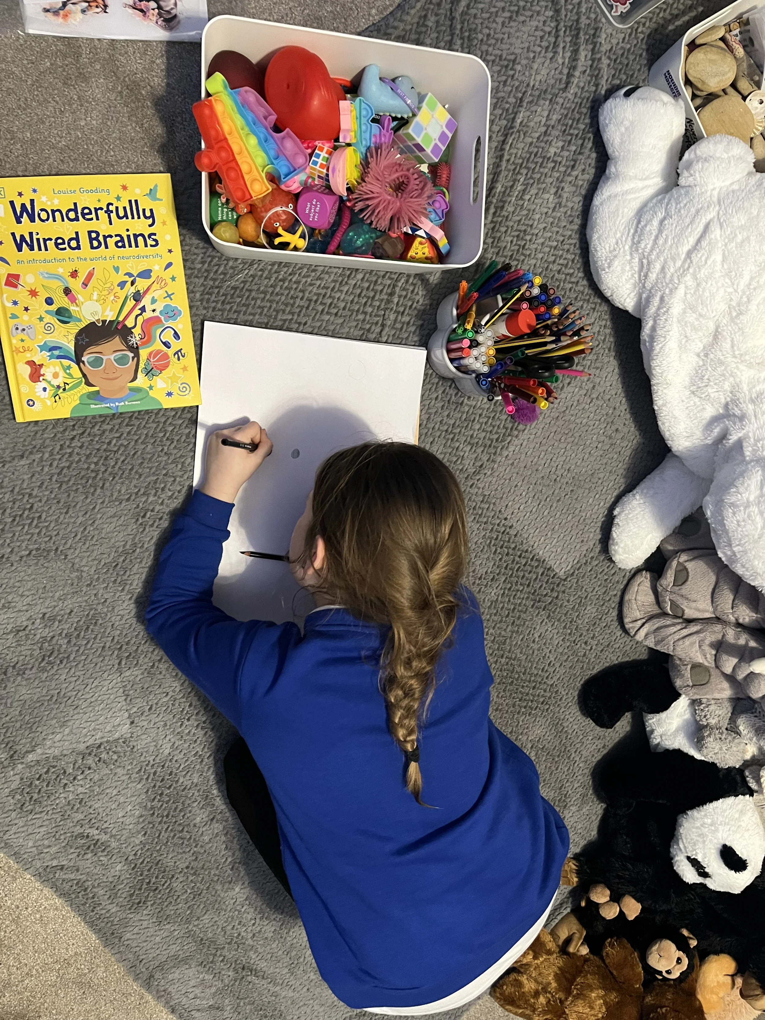 A girl with a plait is sitting on the carpet, drawing on a white sheet of paper. Nearby are books, various toys such as balls, animals, and other small objects, and a cup holding colouring pens.