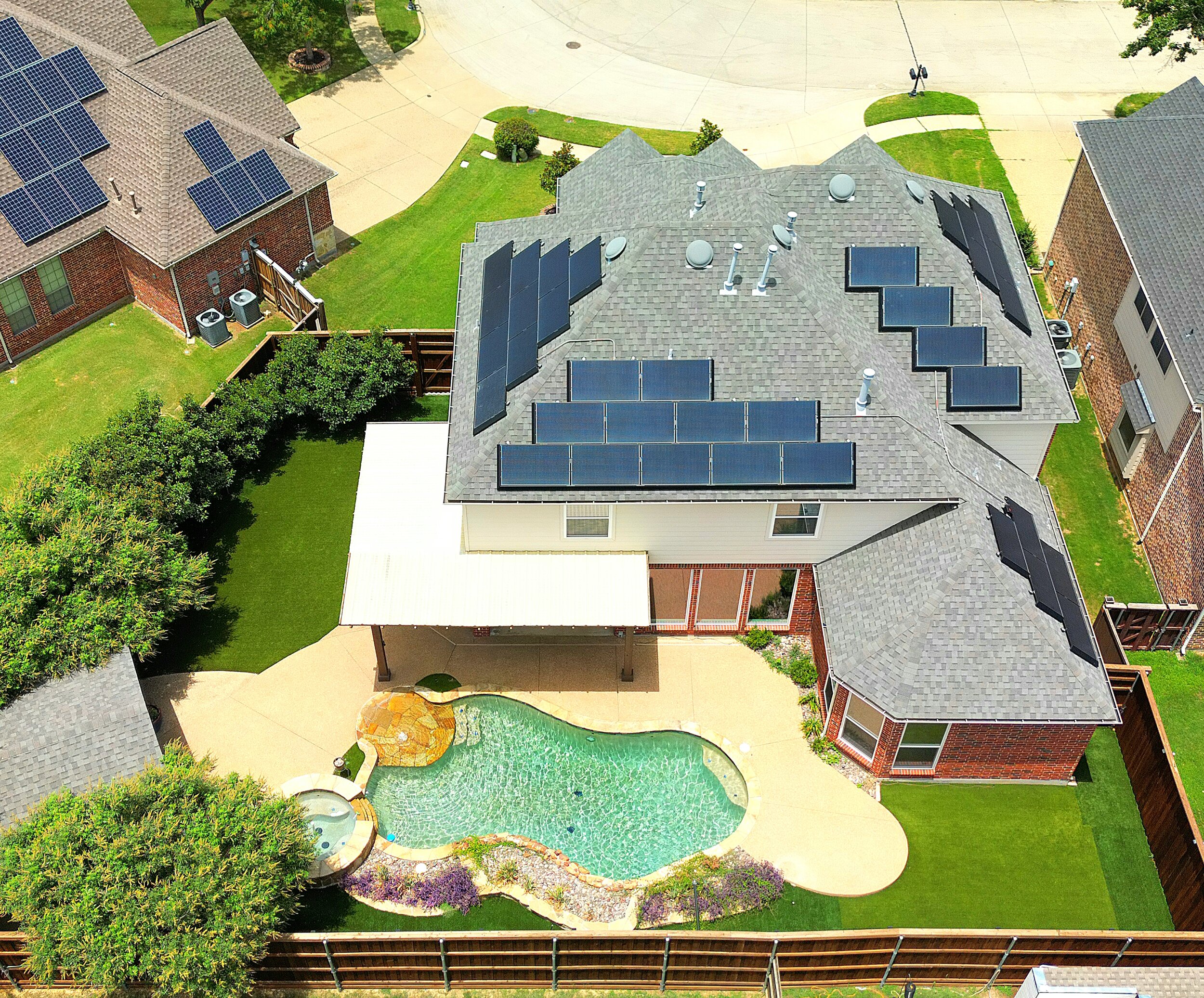 Aerial view of a house with solar panels on the roof, a backyard with a swimming pool, lush green lawn, trees, and a fenced yard, in a suburban neighborhood.