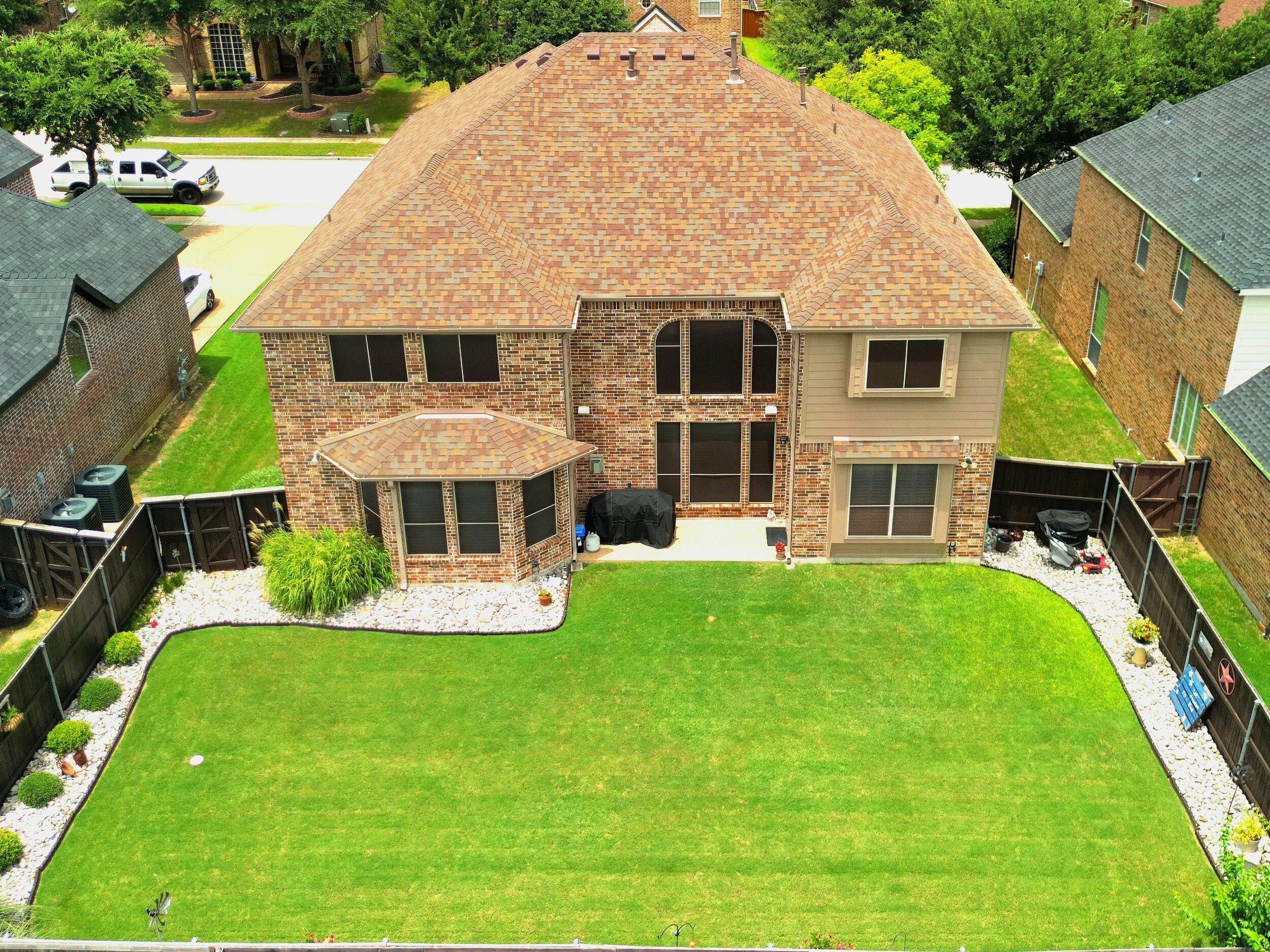 Aerial view of a brick house with a tan roof, backyard with a green lawn, white gravel borders, and fenced yard.