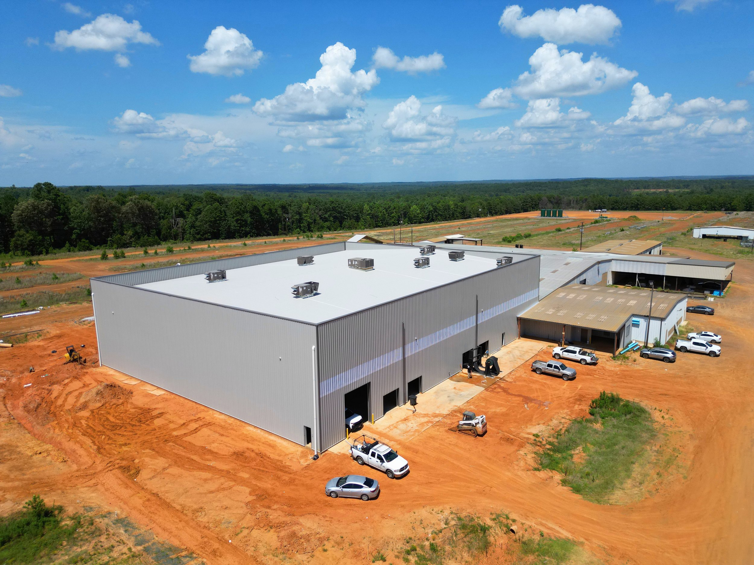A large industrial building under construction with a white roof, surrounded by orange dirt and multiple parked vehicles, set against a green forested landscape with a cloudy blue sky.
